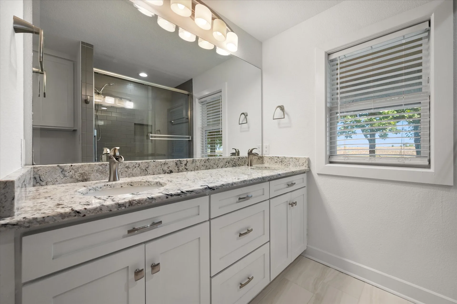 Renovated bathroom with a double vanity, granite counters and a glass-enclosed shower