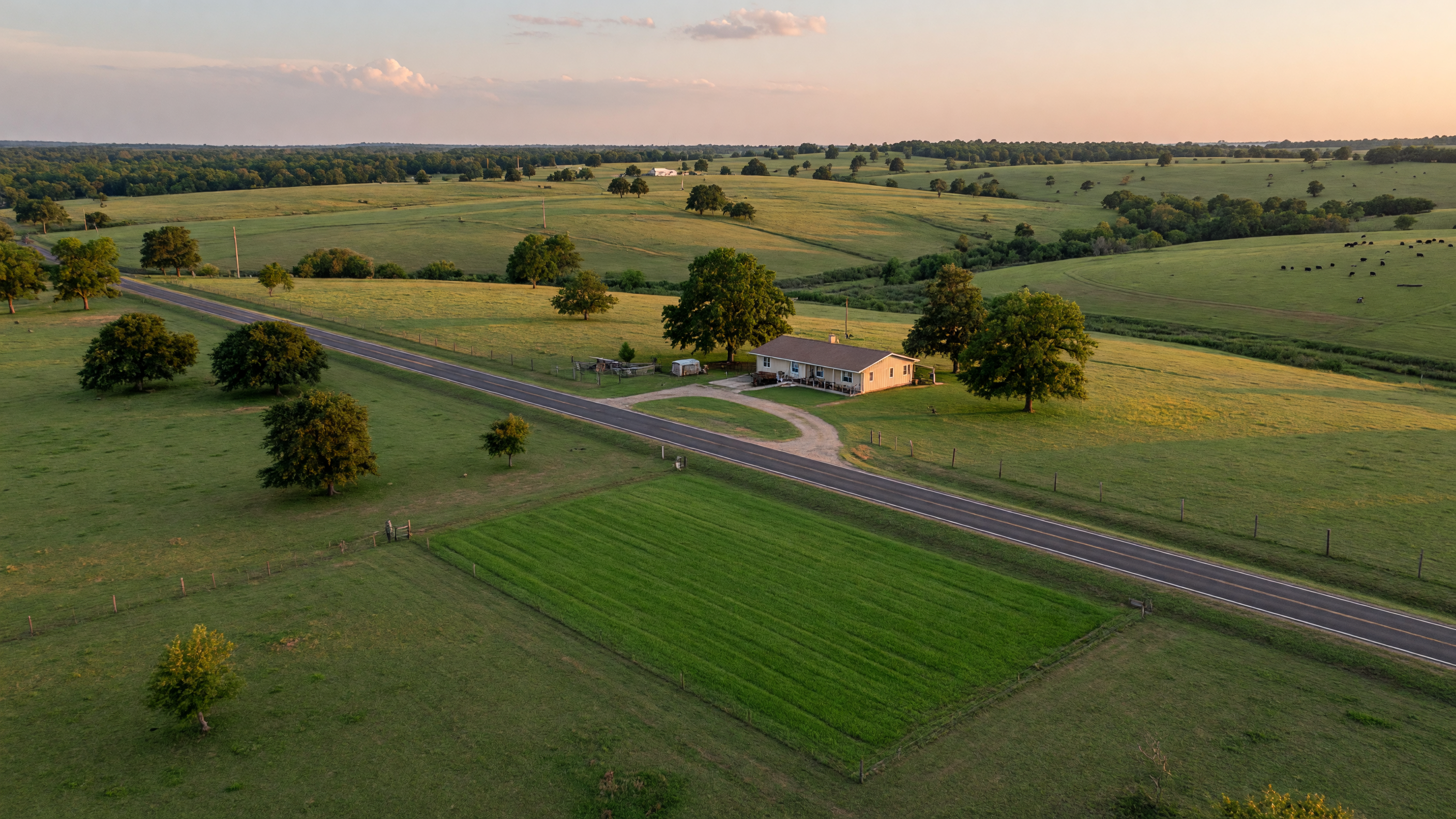 Aerial view of a rural Northeast Texas property with open pastureland, hardwood trees, and a ranch-style home on acreage at golden hour