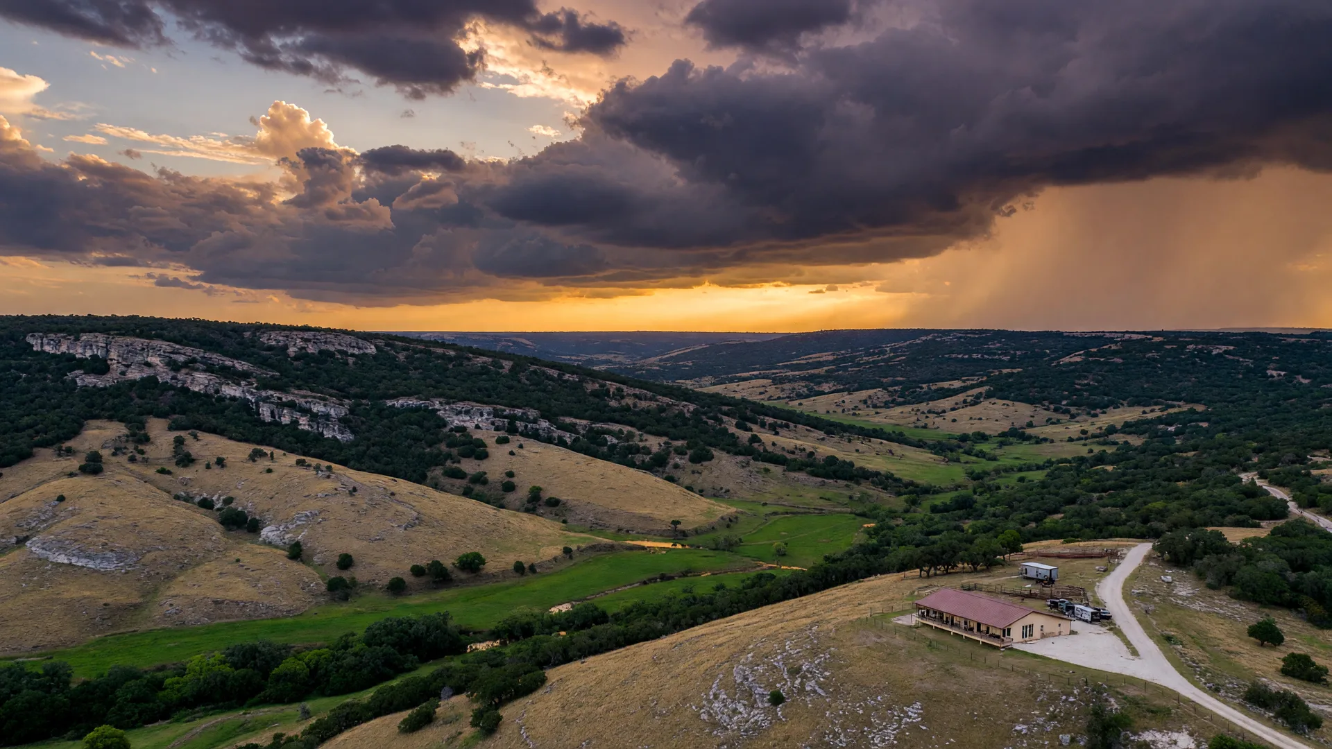 Aerial view of Texas Hill Country terrain showing a dry creek bed winding through limestone hills and live oak groves after a storm, with a residential property visible on elevated ground