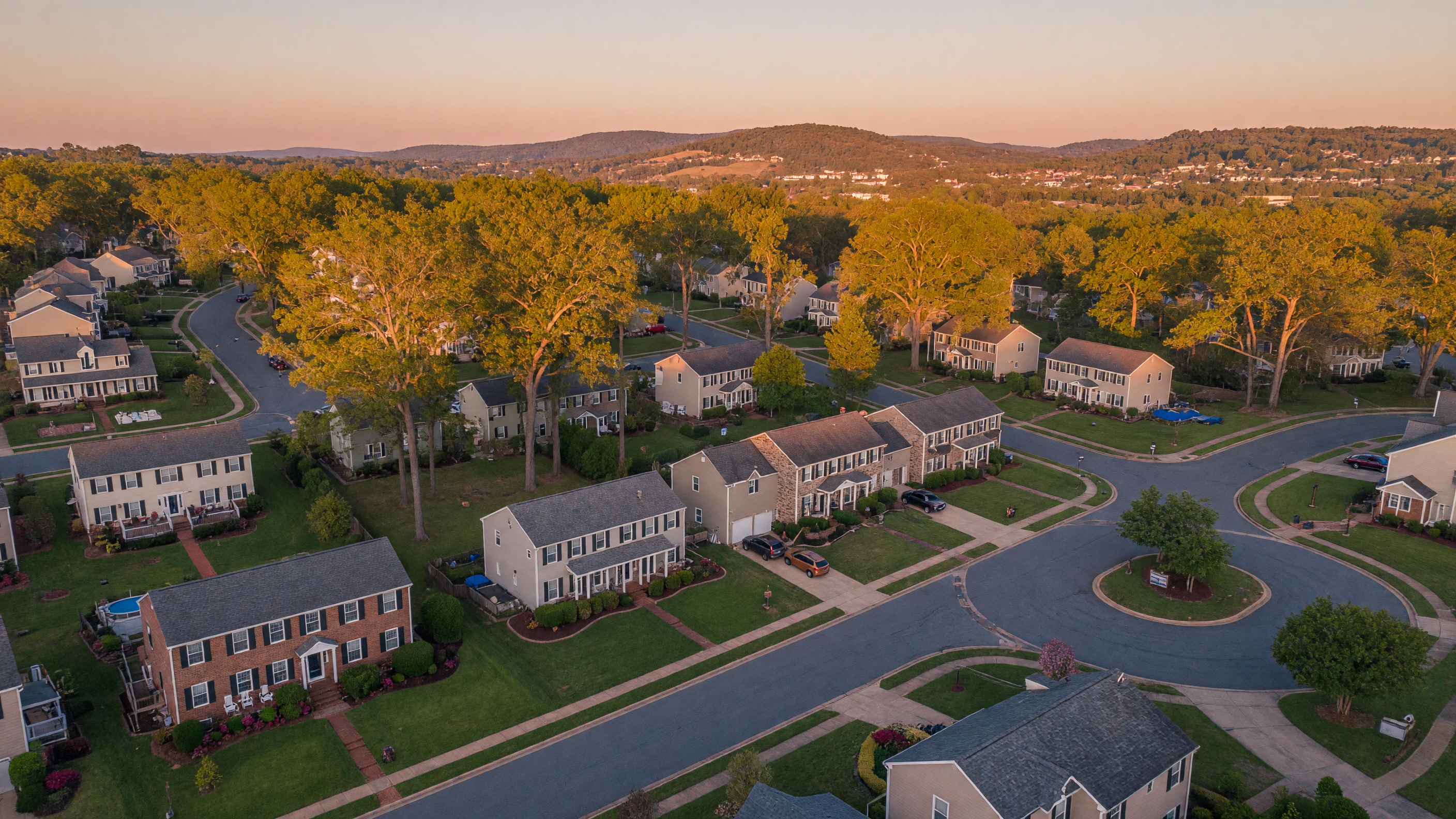 Aerial view of a Virginia suburban neighborhood at golden hour with tree-lined streets, mature landscaping, and rolling hills in the distance