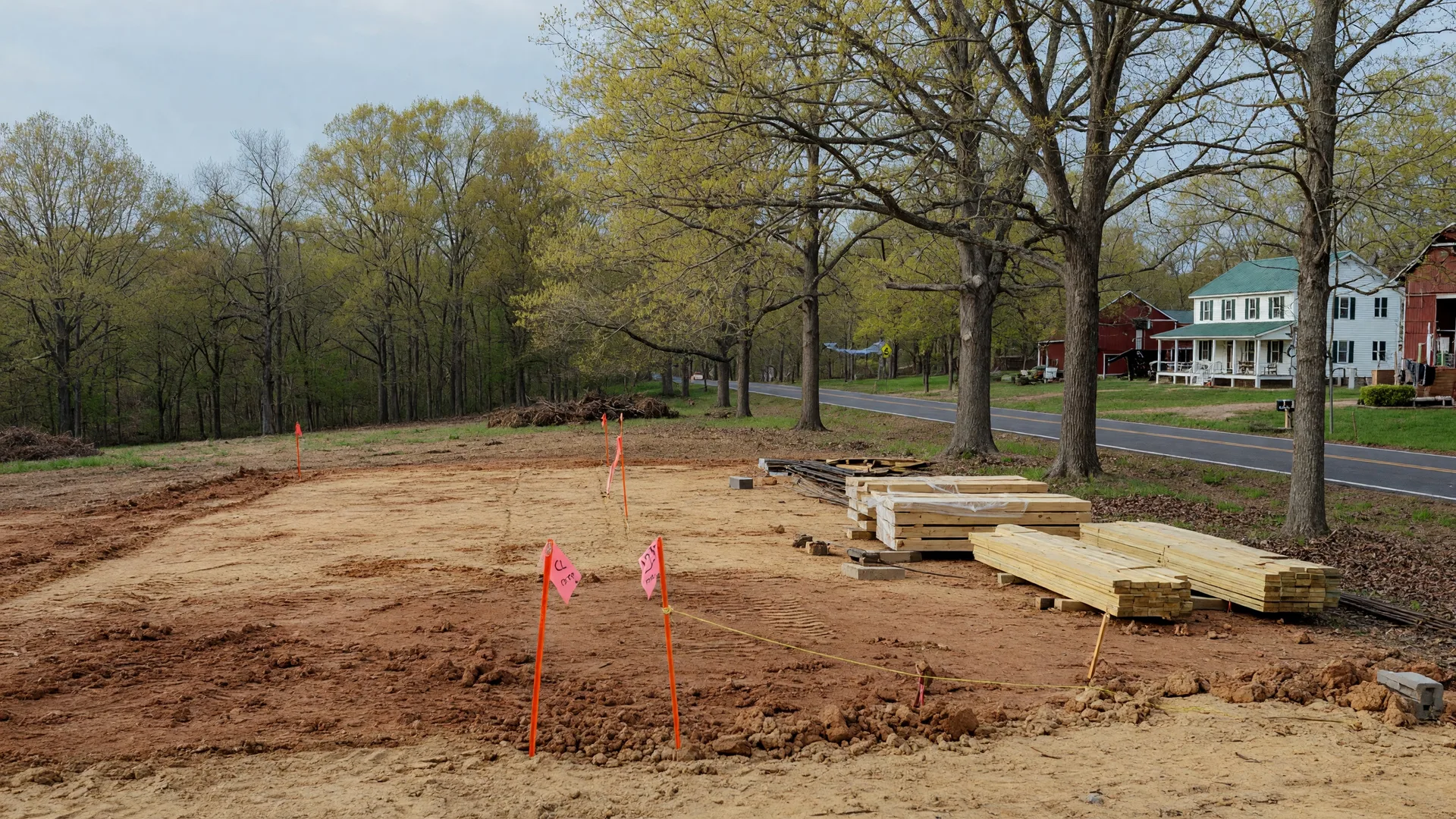 A cleared building site in New Kent County with survey stakes, graded earth, and stacked lumber overlooking farmland and trees under soft afternoon light