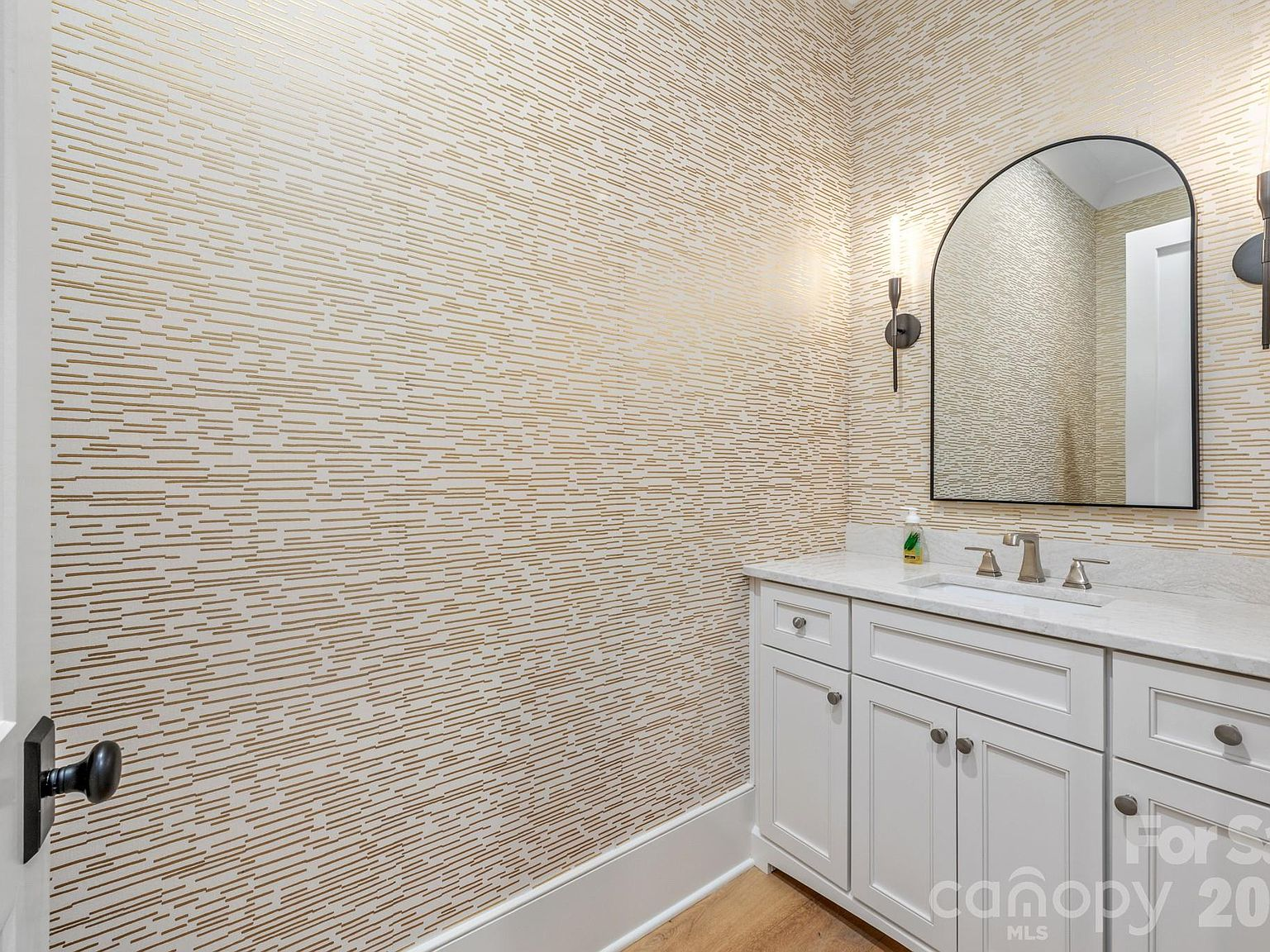 Powder room with gold-patterned wallpaper, white vanity, and arched mirror