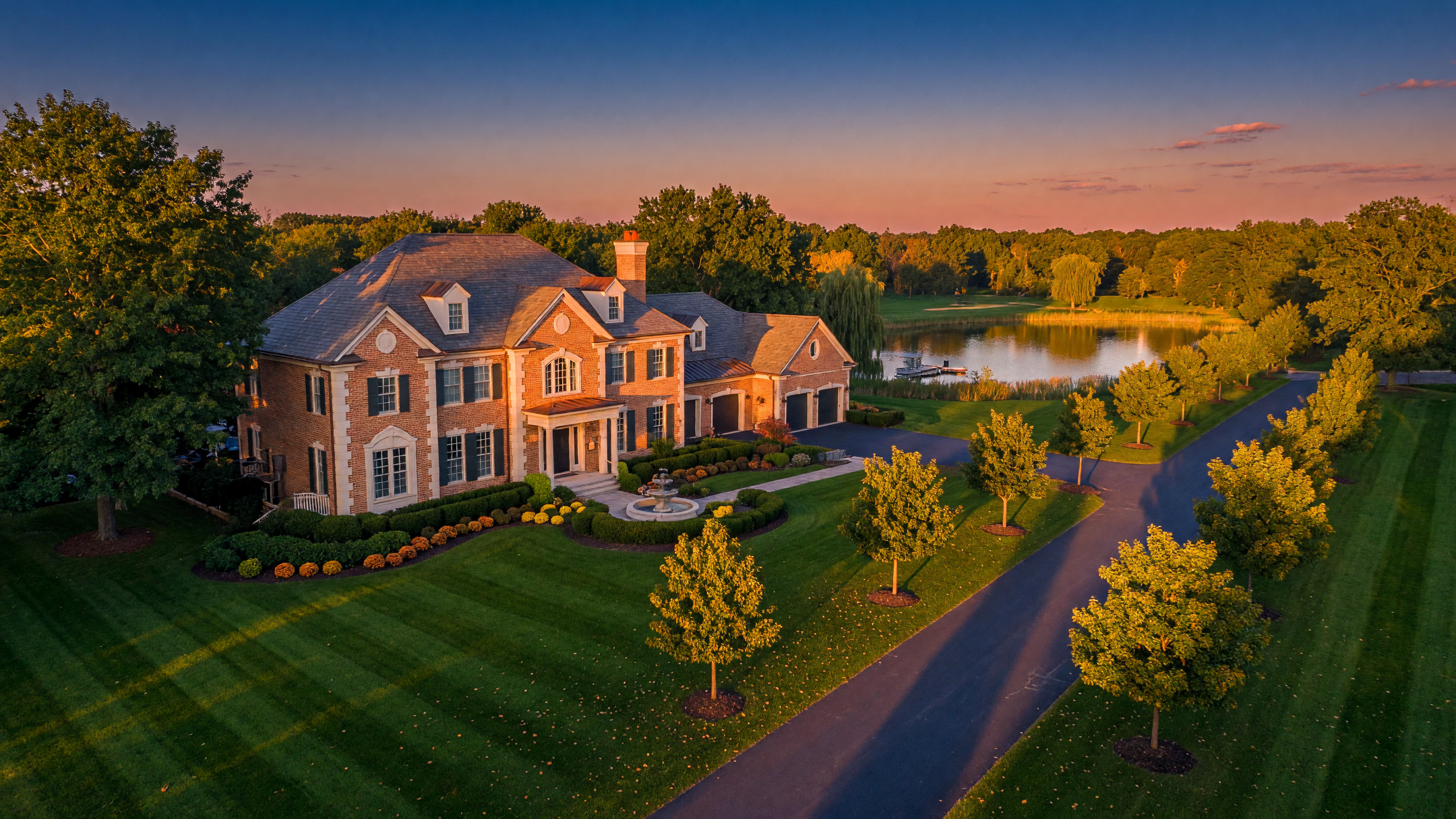 A grand luxury estate home in Mid-Michigan with manicured landscaping, mature trees, and a private pond at golden hour