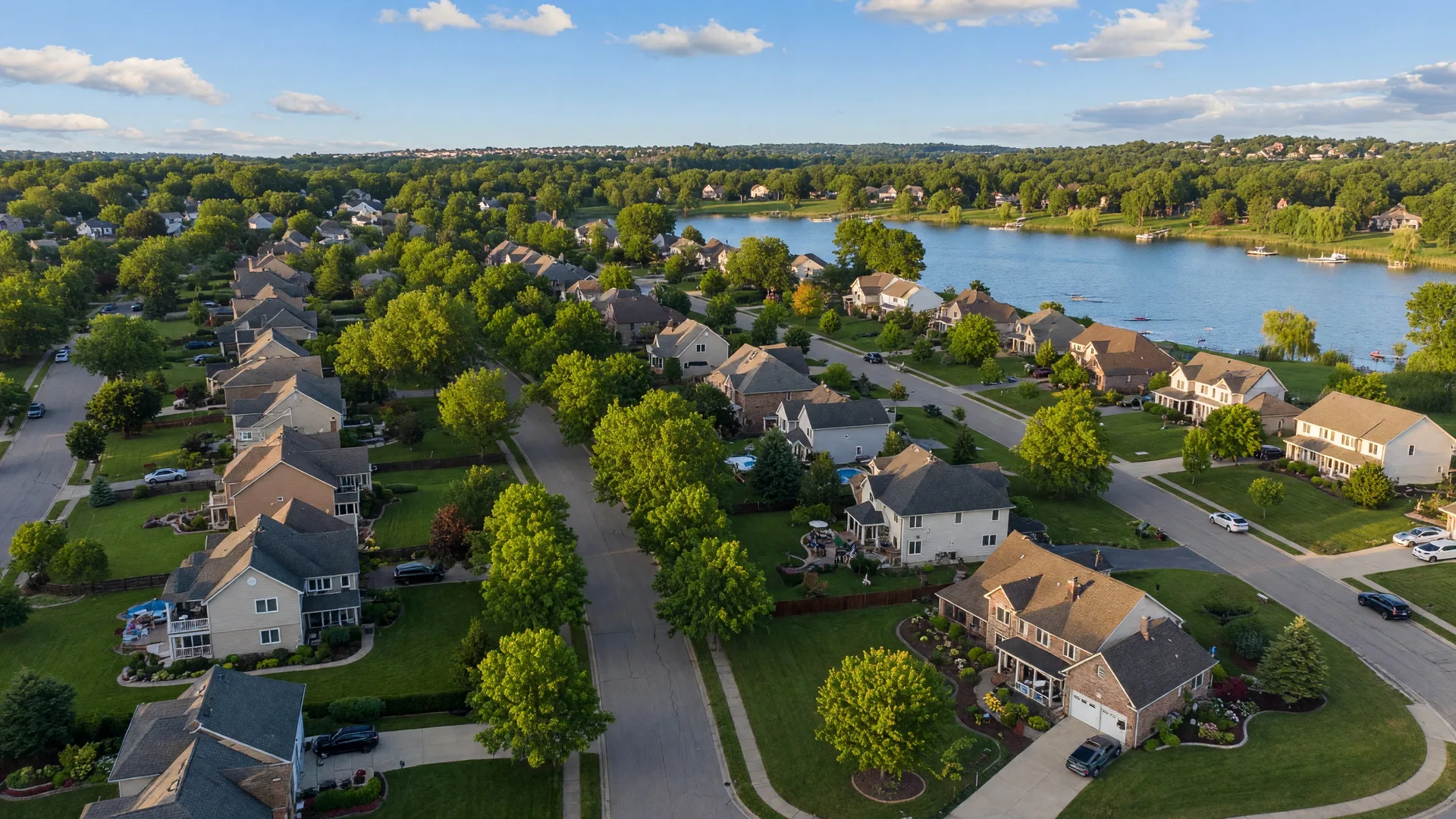 Aerial view of a tree-lined upscale neighborhood in Livingston County Michigan with family homes, green lawns, and a lake in the background on a bright summer day