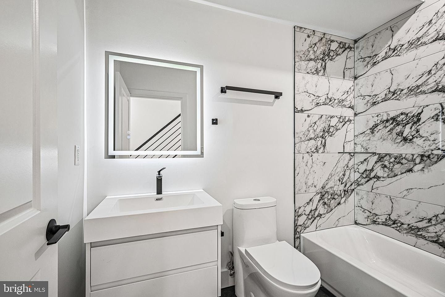 Modern black-and-white marble look tile surrounds the deep bathtub with backlit square mirror