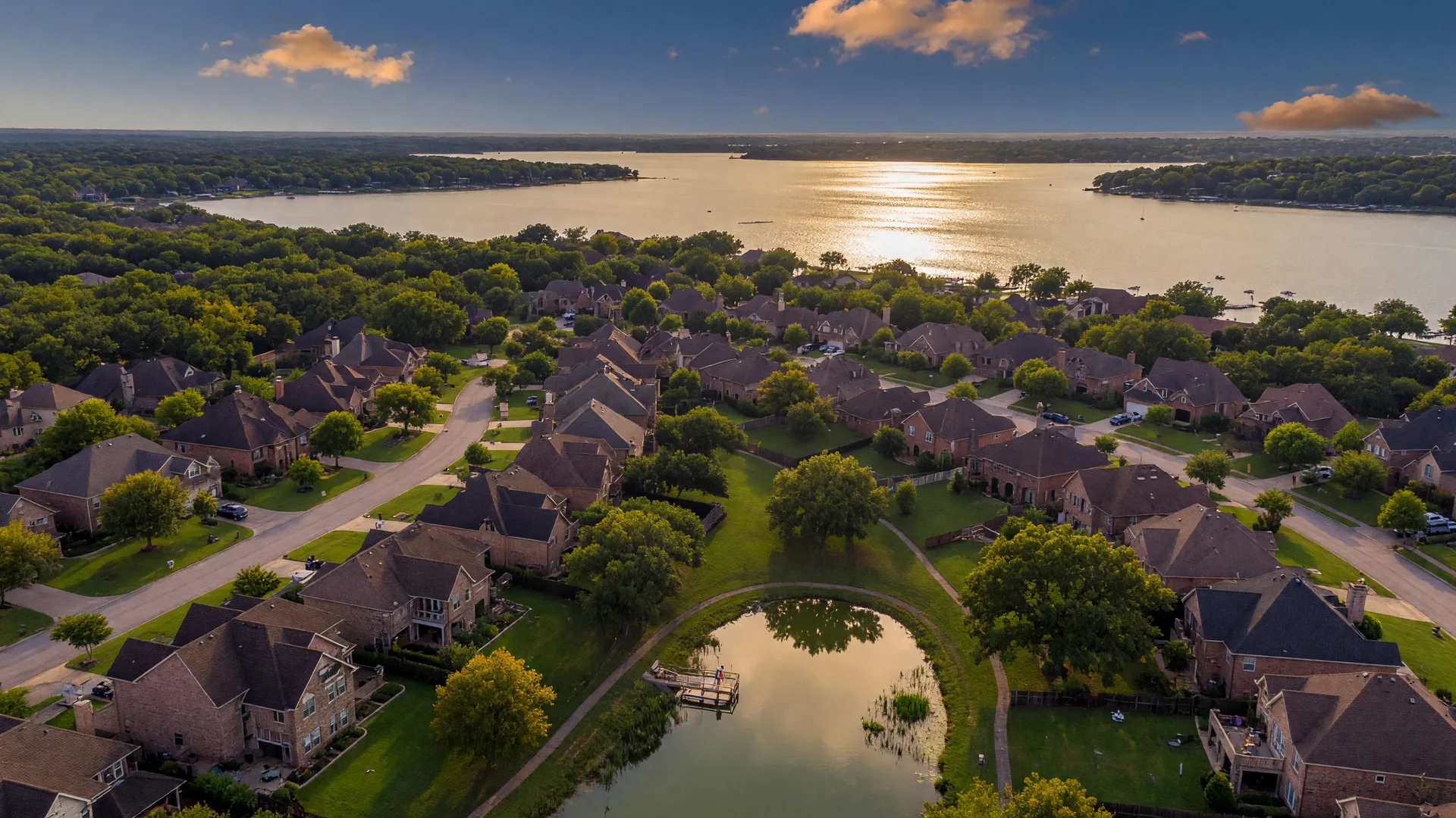 Aerial view of Sycamore Cove neighborhood at golden hour with brick homes, tree-lined streets, and Lake Lewisville in the background