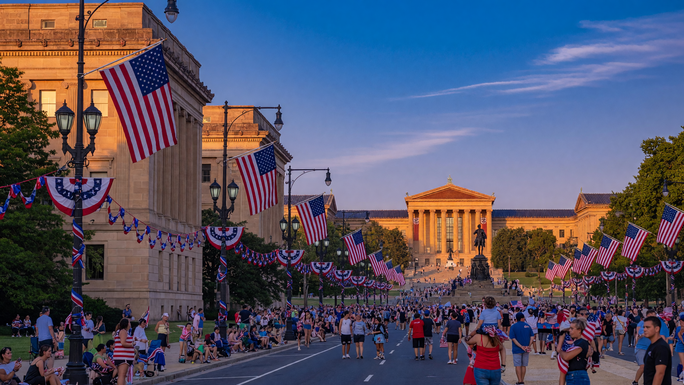 The Benjamin Franklin Parkway in Philadelphia decorated with American flags and patriotic bunting for a summer celebration, with crowds gathering toward the Art Museum