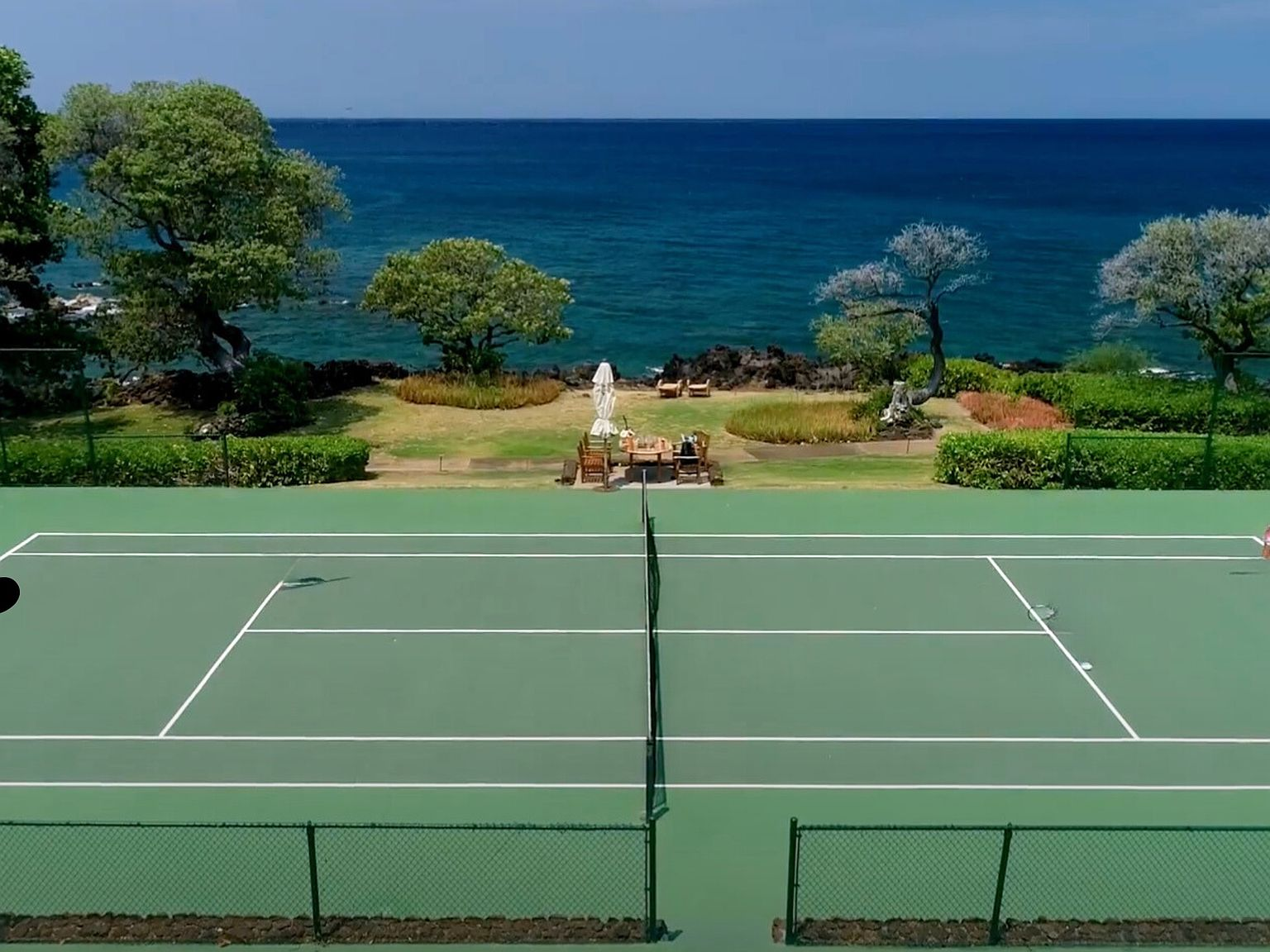 Lush oceanfront grass tennis court surrounded by coconut palms and basalt rock shores