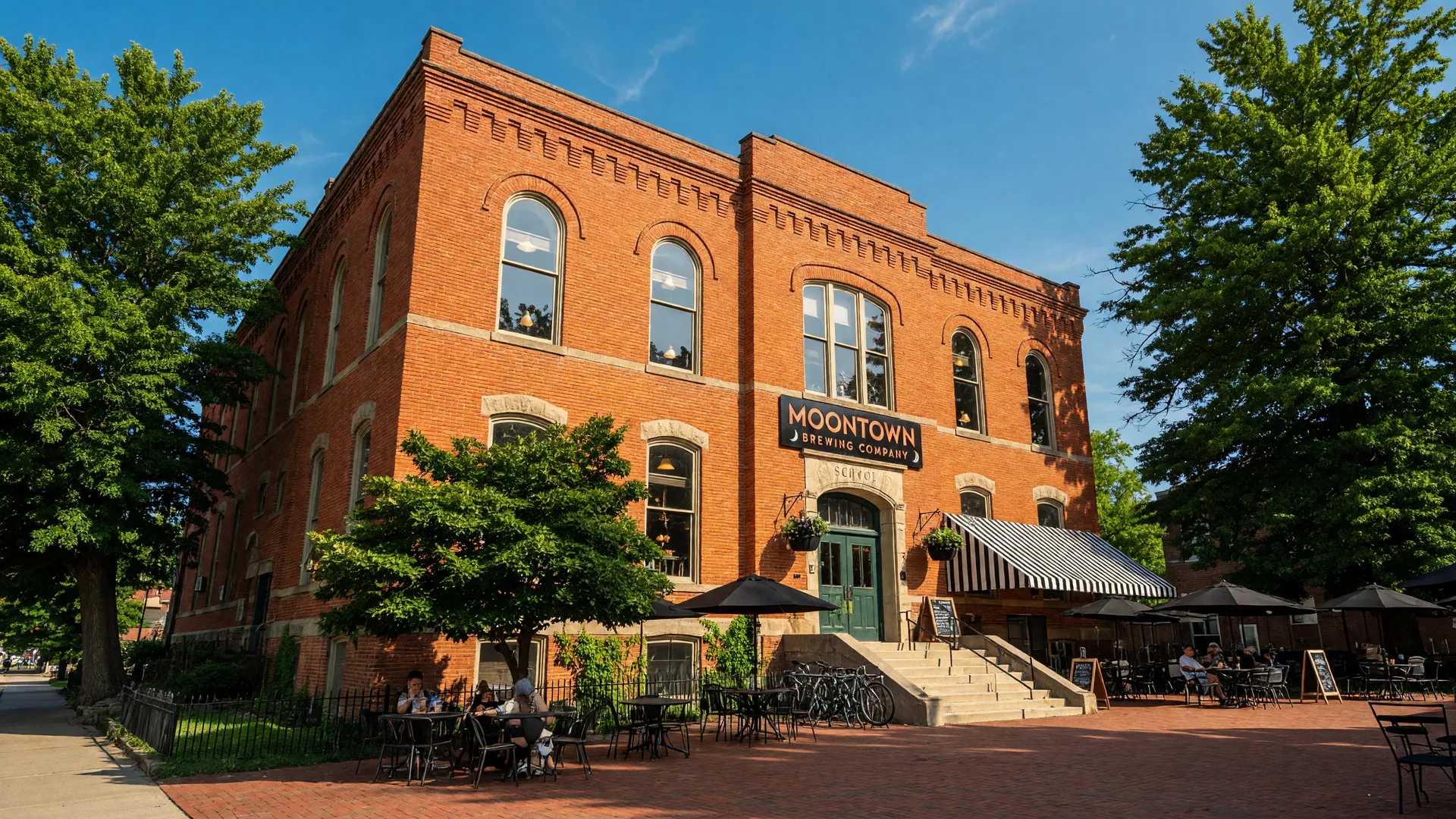 Moontown Brewing Company in Whitestown Indiana — a beautifully restored historic school building with red brick exterior, arched windows, and a welcoming brewery patio on a summer afternoon