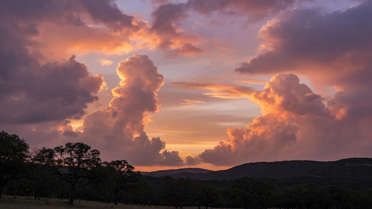 A dramatic Hill Country sunset sky with towering cumulus clouds lit in warm orange and gold above a silhouette of live oak trees — the vast Texas sky is a daily feature