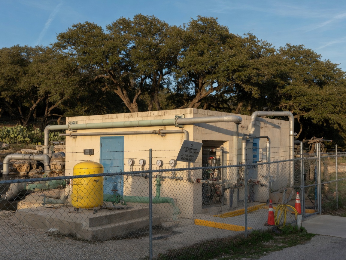 New residential infrastructure under construction in the Texas Hill Country, showing a freshly paved road with concrete curbing, utility pedestals, and graded lots with preserved oak trees along the corridor