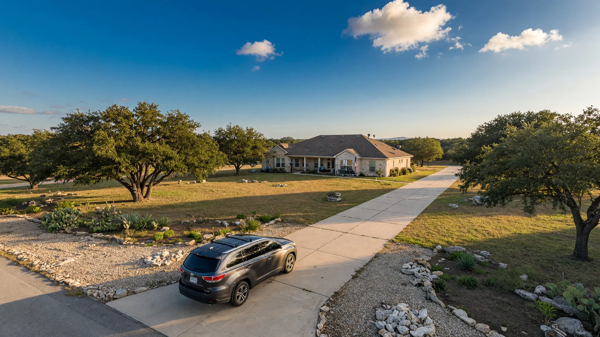 A long concrete driveway leading to a Texas Hill Country home set far back from the road, with mature live oak trees and a wide blue sky overhead