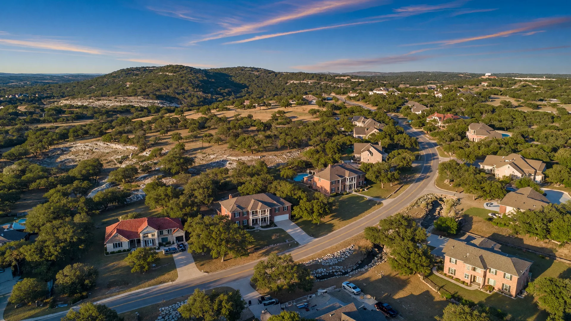 Aerial view of a residential Texas Hill Country neighborhood in Boerne with limestone homes, live oak trees, and rolling hills at golden hour