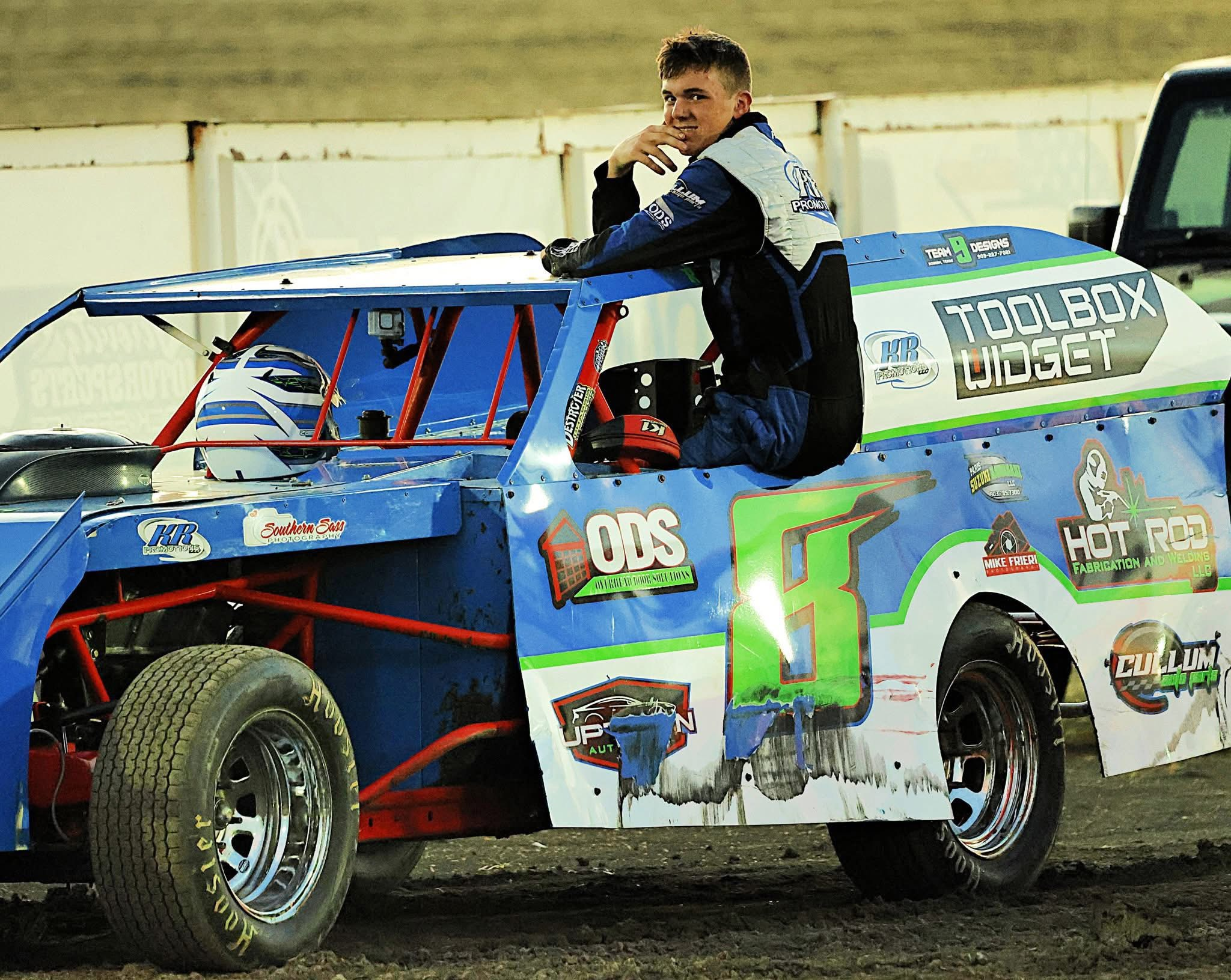 Young race car driver sitting on the door of a blue and white stock car in the paddock
