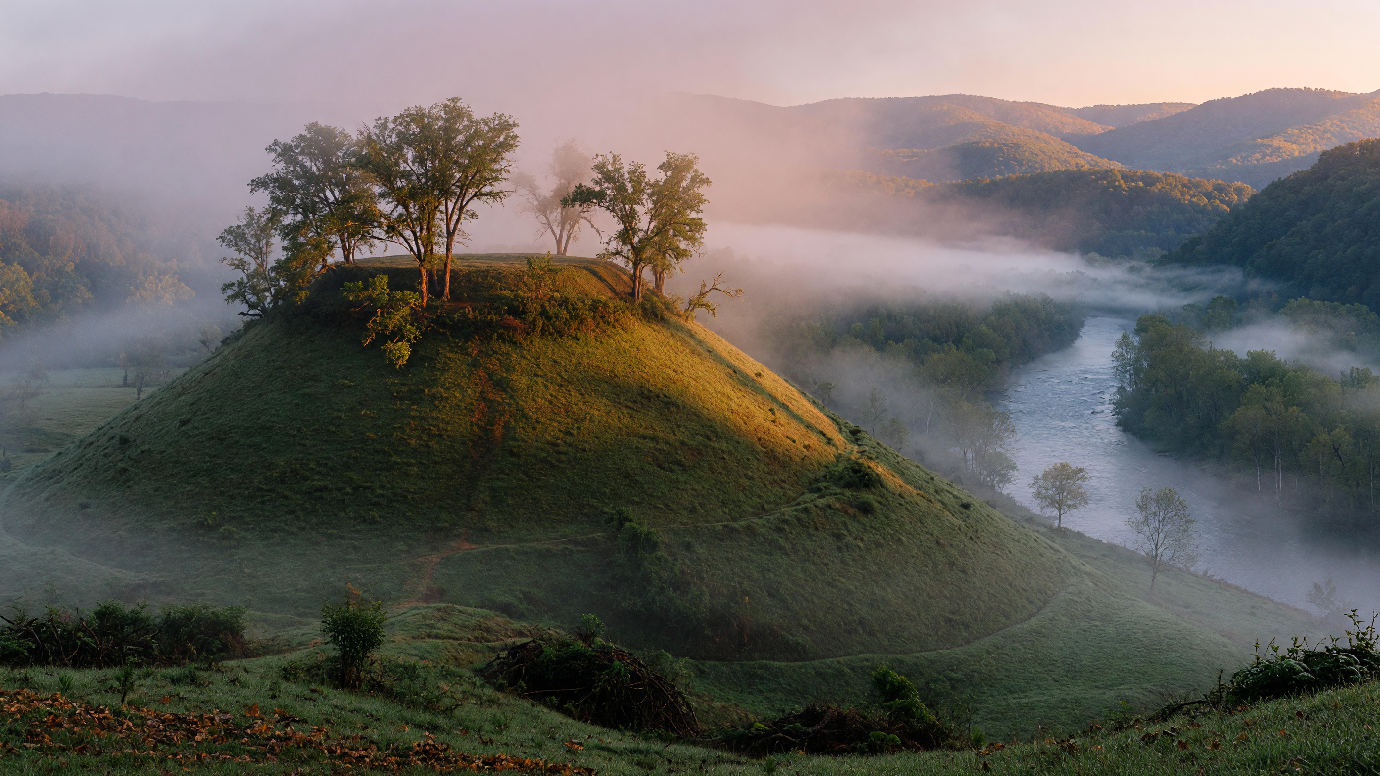 An ancient grass-covered mound rising above a misty river valley at dawn, surrounded by mature hardwood trees in the Appalachian foothills, evoking the Cherokee settlements that once occupied the Tugaloo Valley