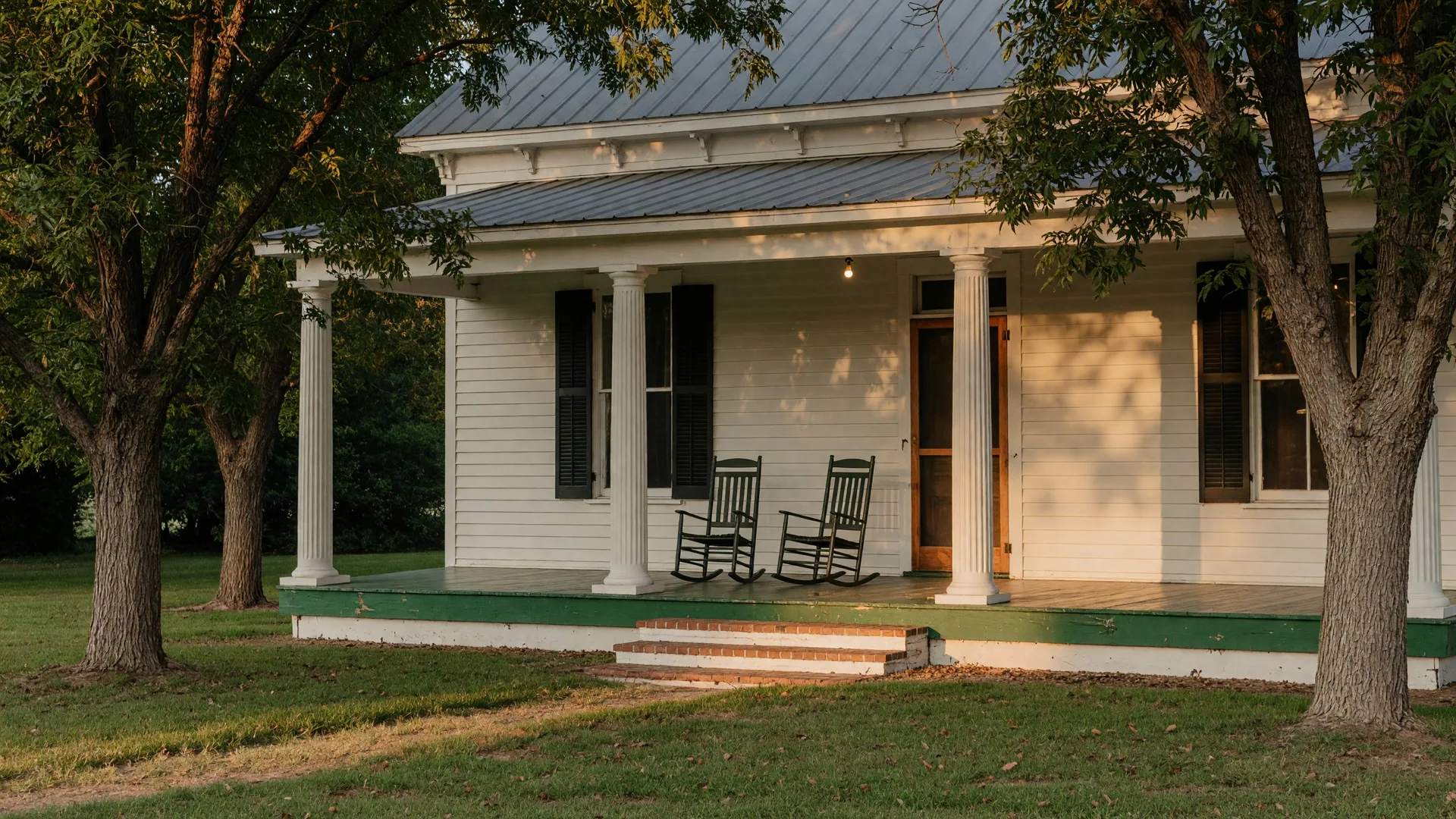 Historic farmhouse porch with white columns, rocking chairs, and mature pecan trees casting dappled shade across the front lawn