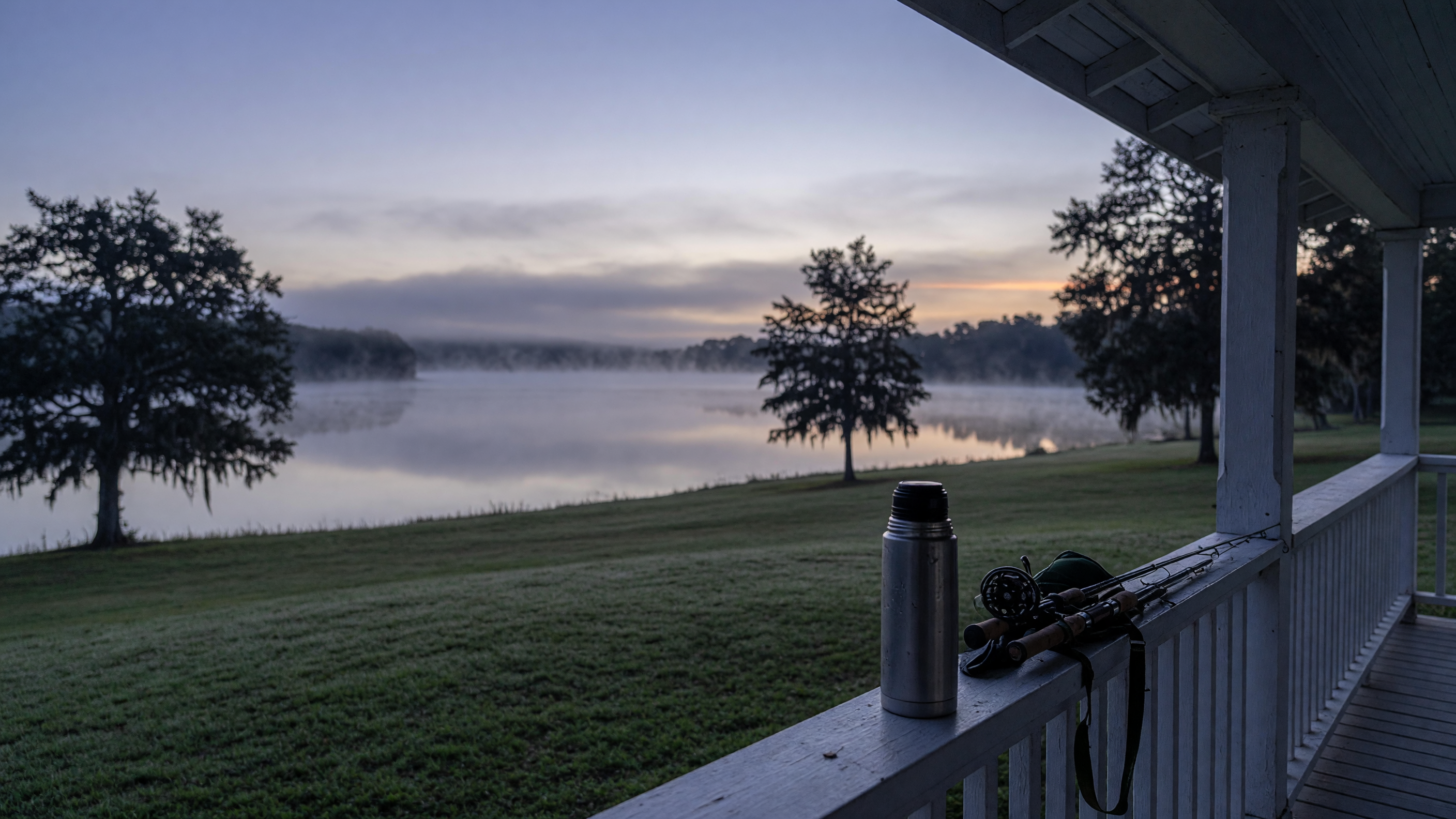 Pre-dawn view from a covered porch overlooking a calm lake with mist rising off the water, a coffee thermos resting on the rail