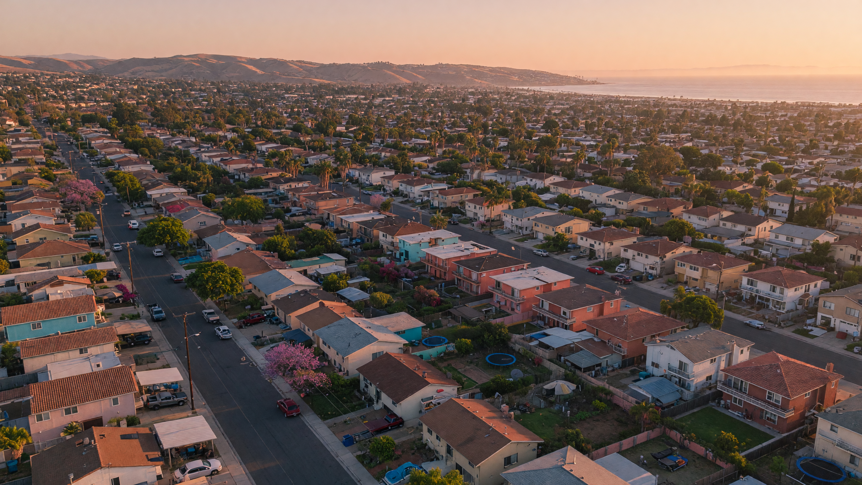 Aerial view of a diverse San Diego County residential neighborhood at golden hour, showing a mix of single-family homes and small apartment buildings stretching toward distant hills