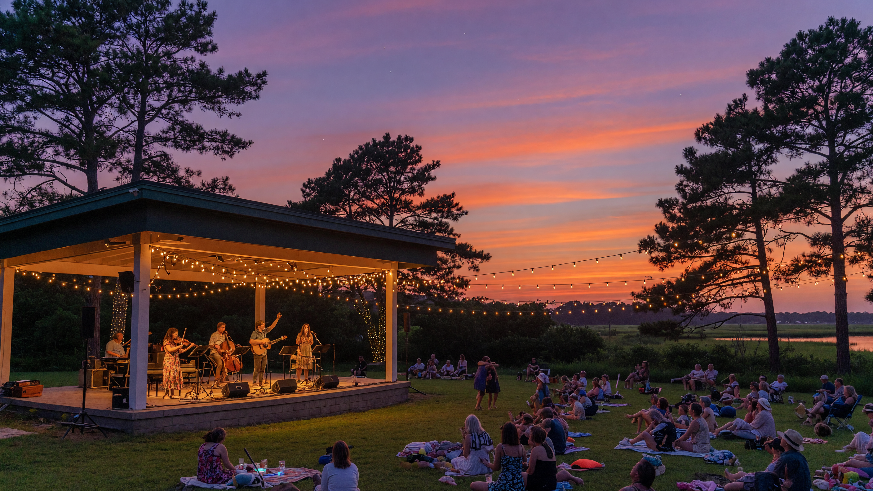 Outdoor summer concert at the Freeman Arts Pavilion at twilight with warm string lights over the stage
