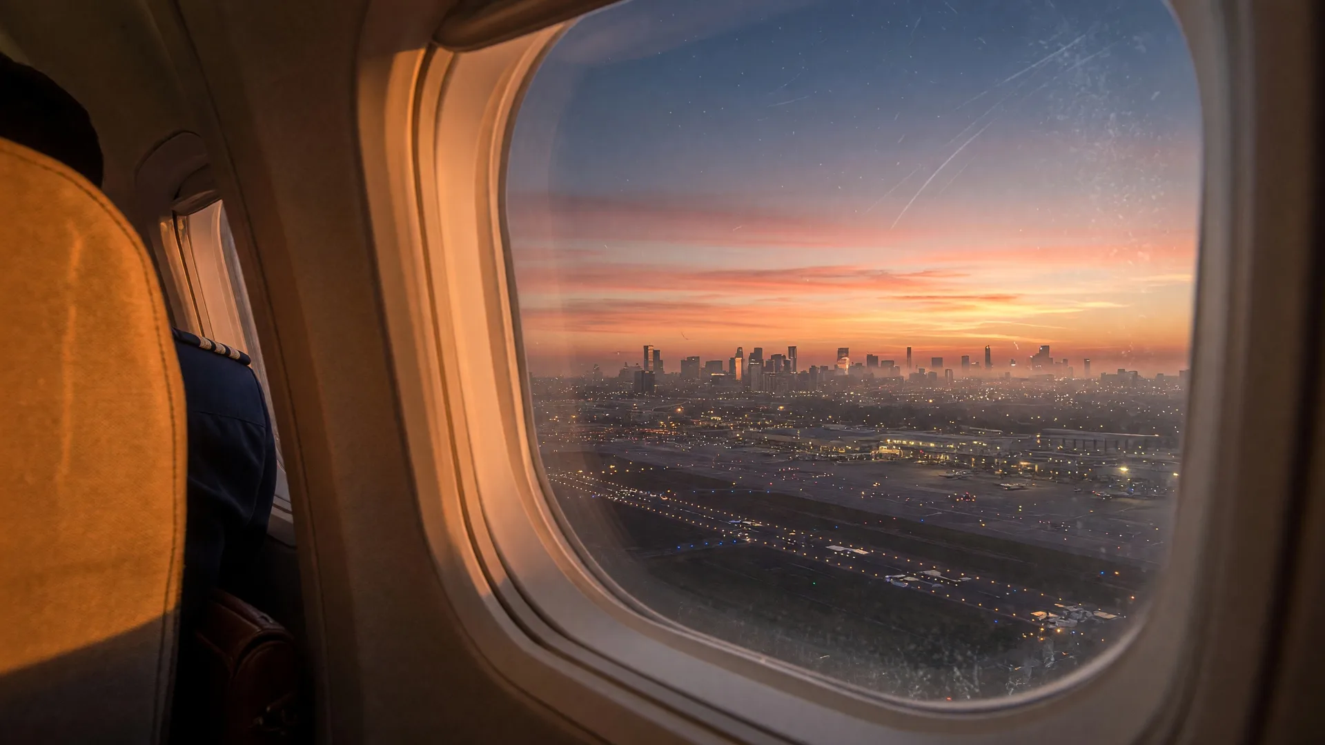 View from an airplane window at sunrise showing a city skyline and runway lights below