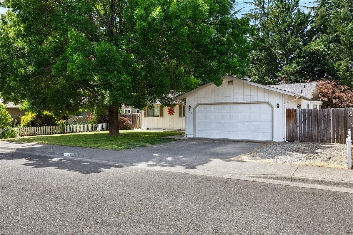 Front exterior of 1212 SW Cottonwood St, a single-story ranch home with white board-and-batten siding, two-car garage, and mature shade trees