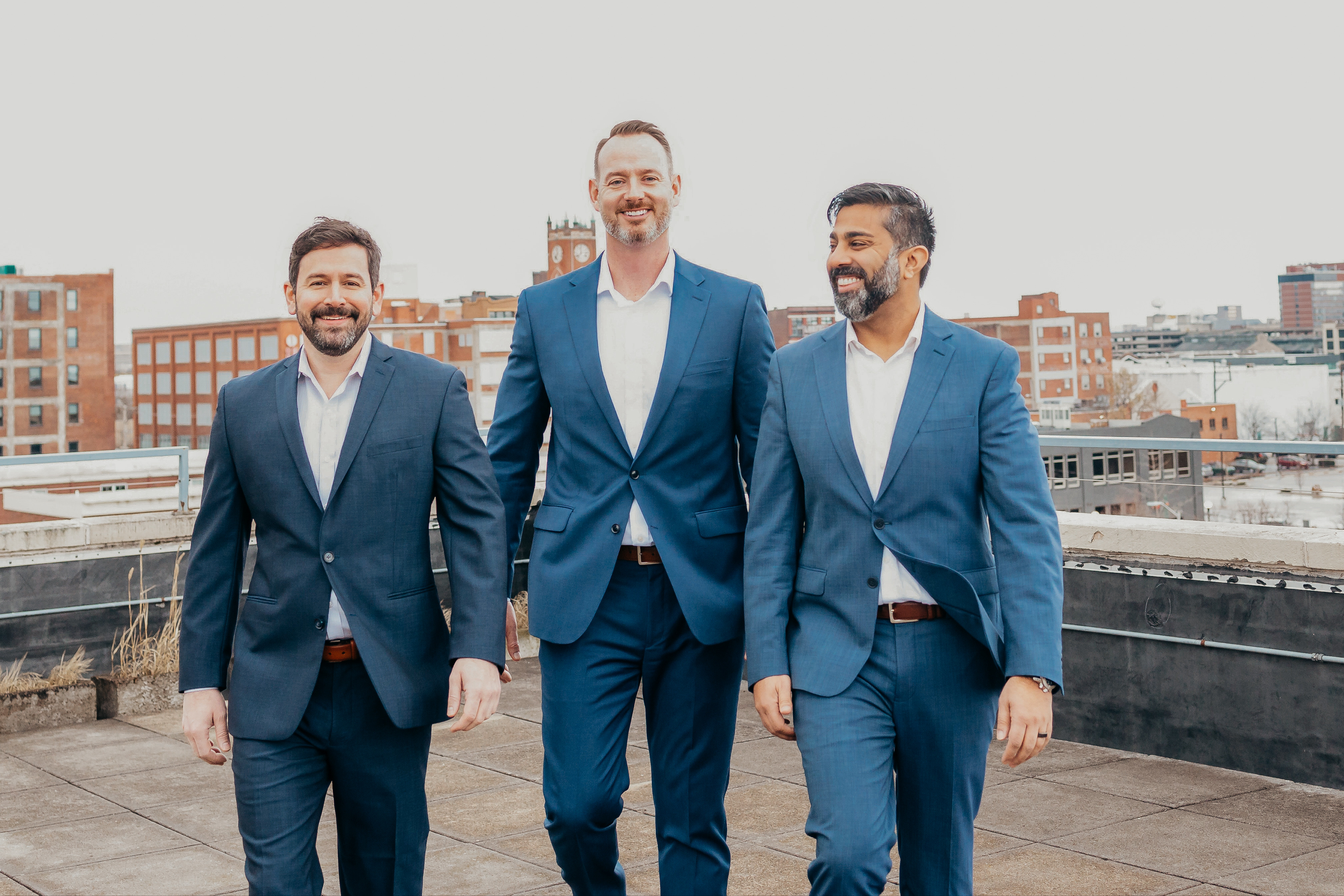 Three men in formal navy suits walking together on an urban rooftop with a blurred city skyline background
