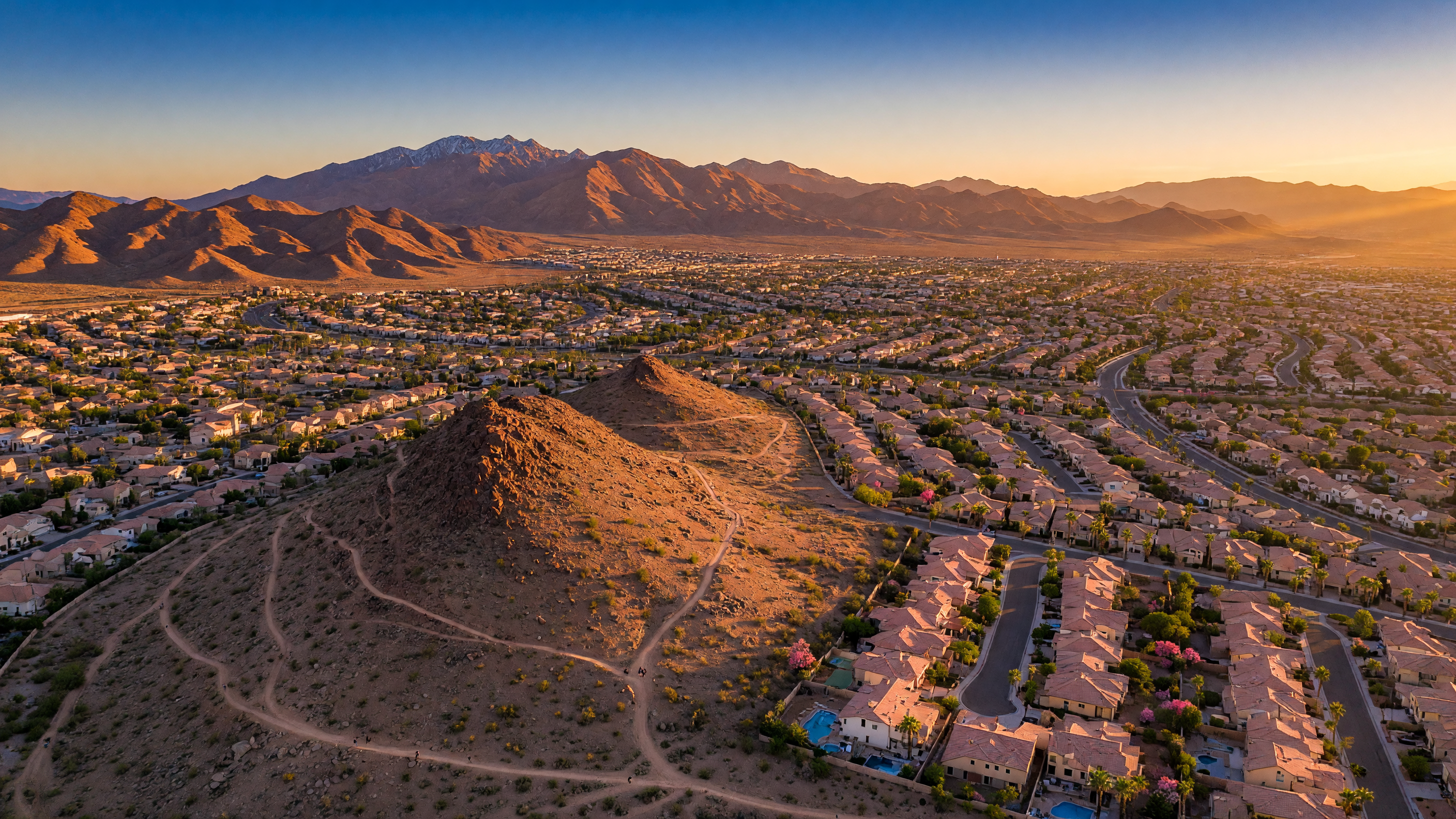 Aerial view of the Mountain's Edge master-planned community in Southwest Las Vegas showing Exploration Peak, neighborhoods with palm-tree-lined streets, parks, and the Spring Mountains at golden hour