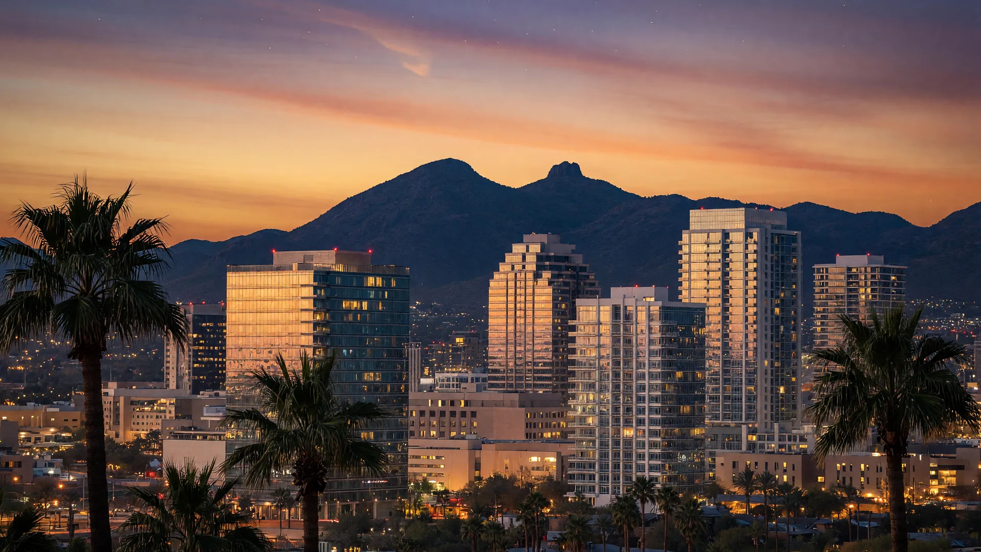 Phoenix downtown skyline at dusk with Cityscape buildings, palm trees in the foreground, and Camelback Mountain silhouette behind under golden-hour light