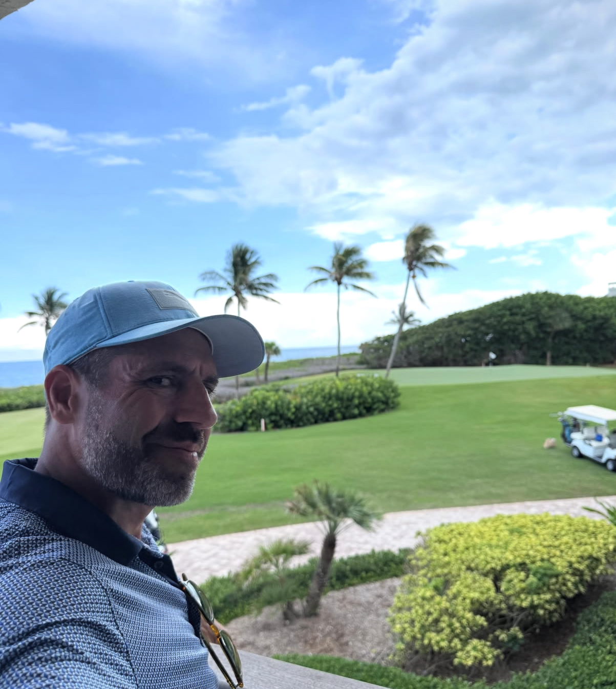 Alex Haigh on a balcony overlooking a coastal golf course with ocean and palm trees