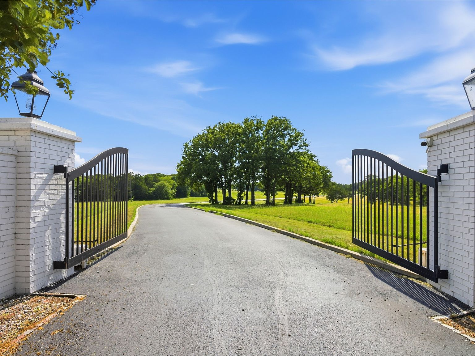 Grand gated entry to 1073 Gordonville Rd, white brick monument pillars with lantern fixtures frame a paved curving driveway lined by mature oak trees leading into 50 acres of rolling Texas countryside
