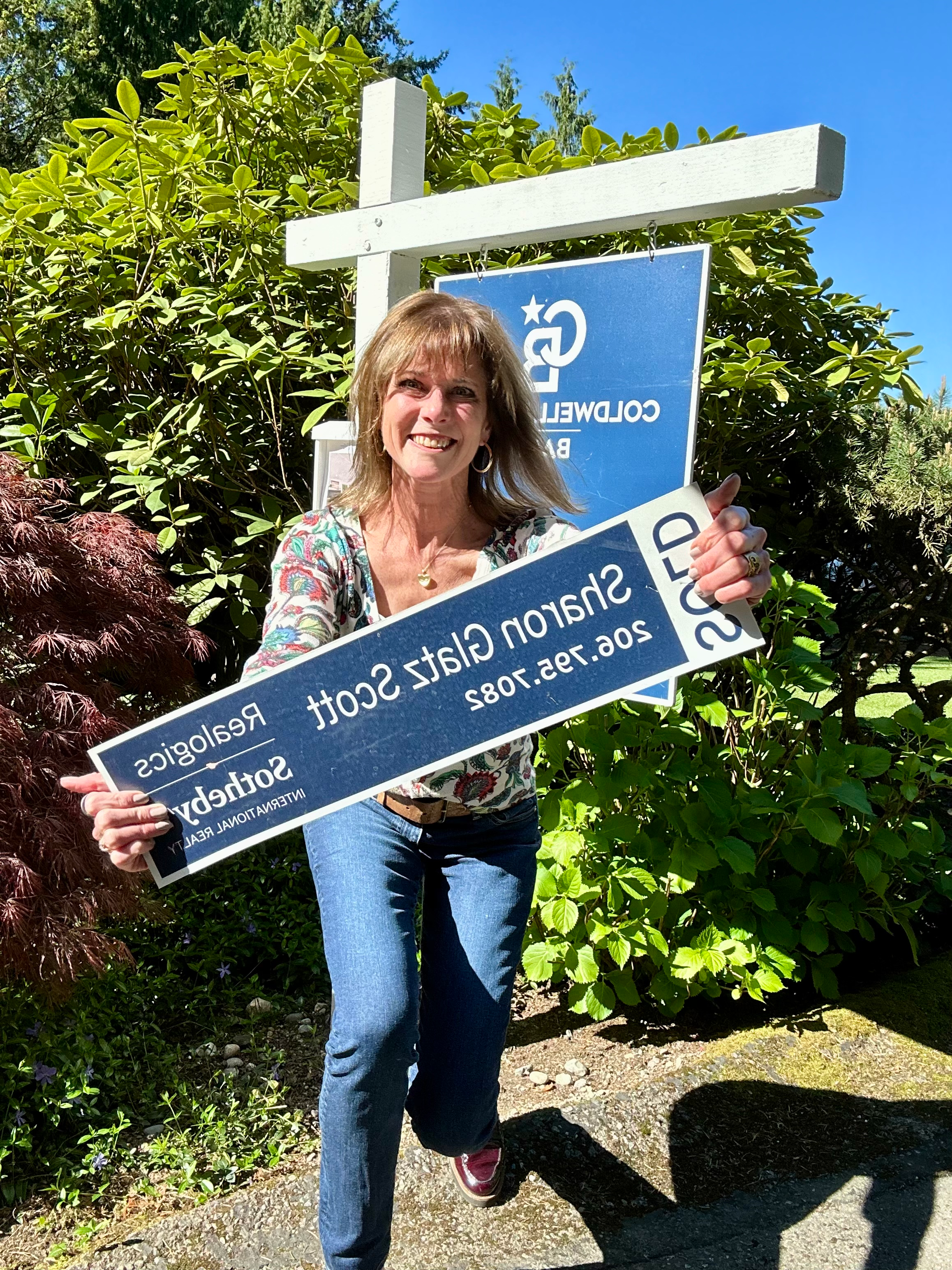 Sharon Glatz Scott holds a Sold sign in front of a real estate post outdoors