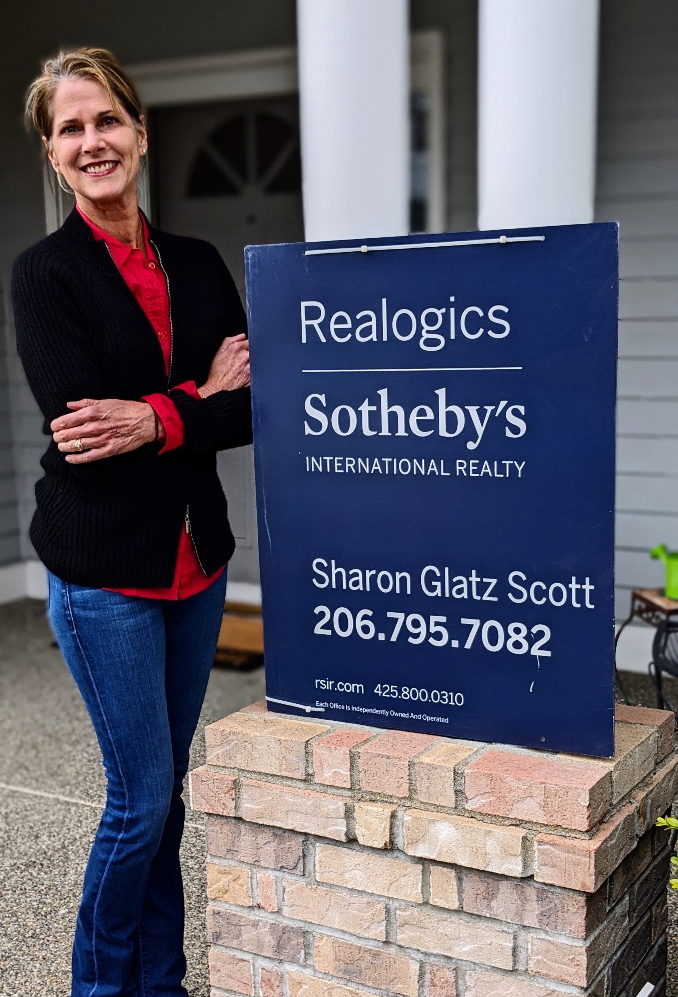 Sharon Glatz Scott stands smiling with her arms crossed next to a branded Realogics Sotheby's International Realty yard sign