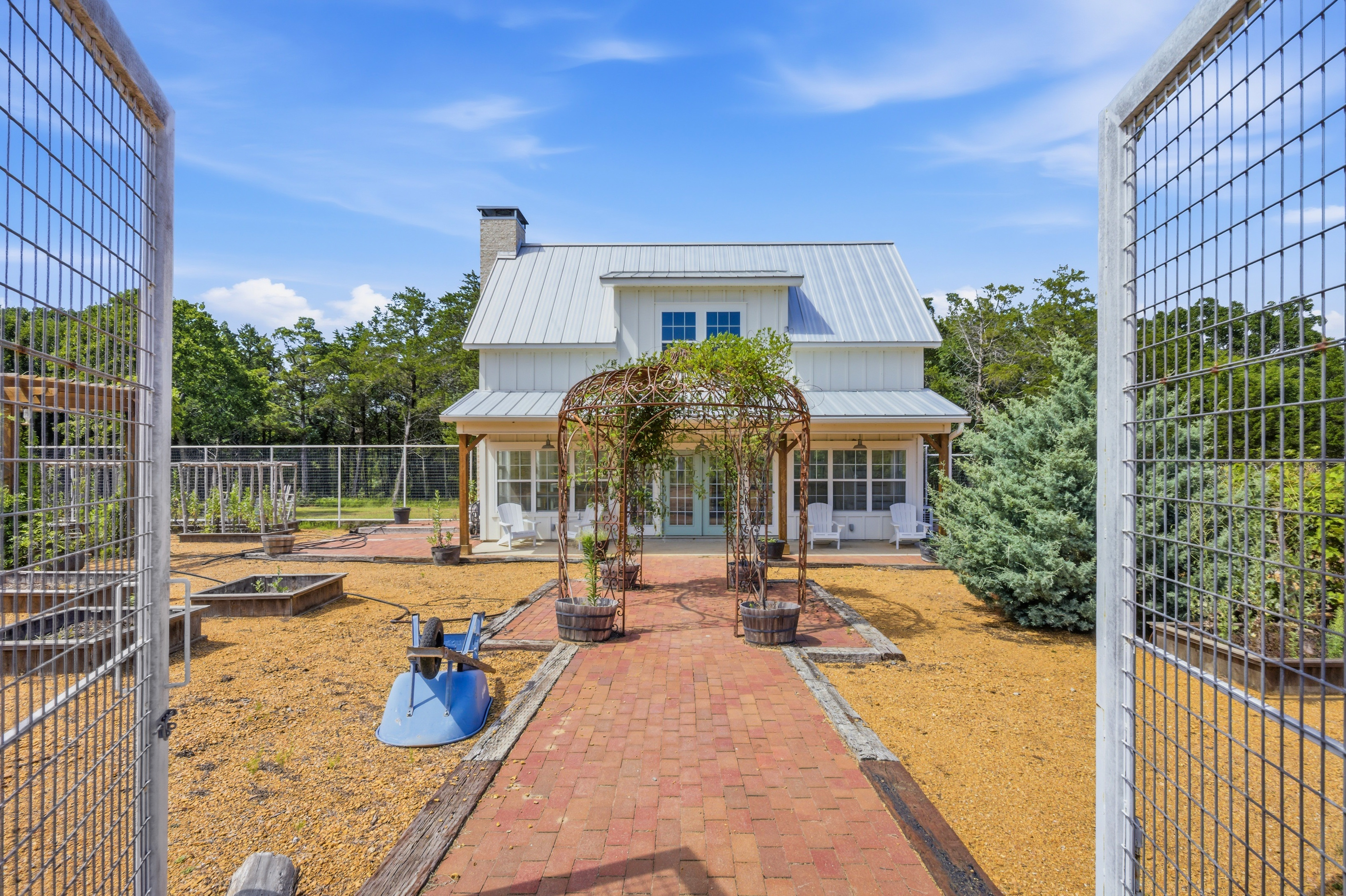 Ground-level view through an open gate into the fenced greenhouse garden at 1073 Gordonville Rd, a brick pathway leads past a decorative metal arbor toward raised garden beds and the two-story guest cottage