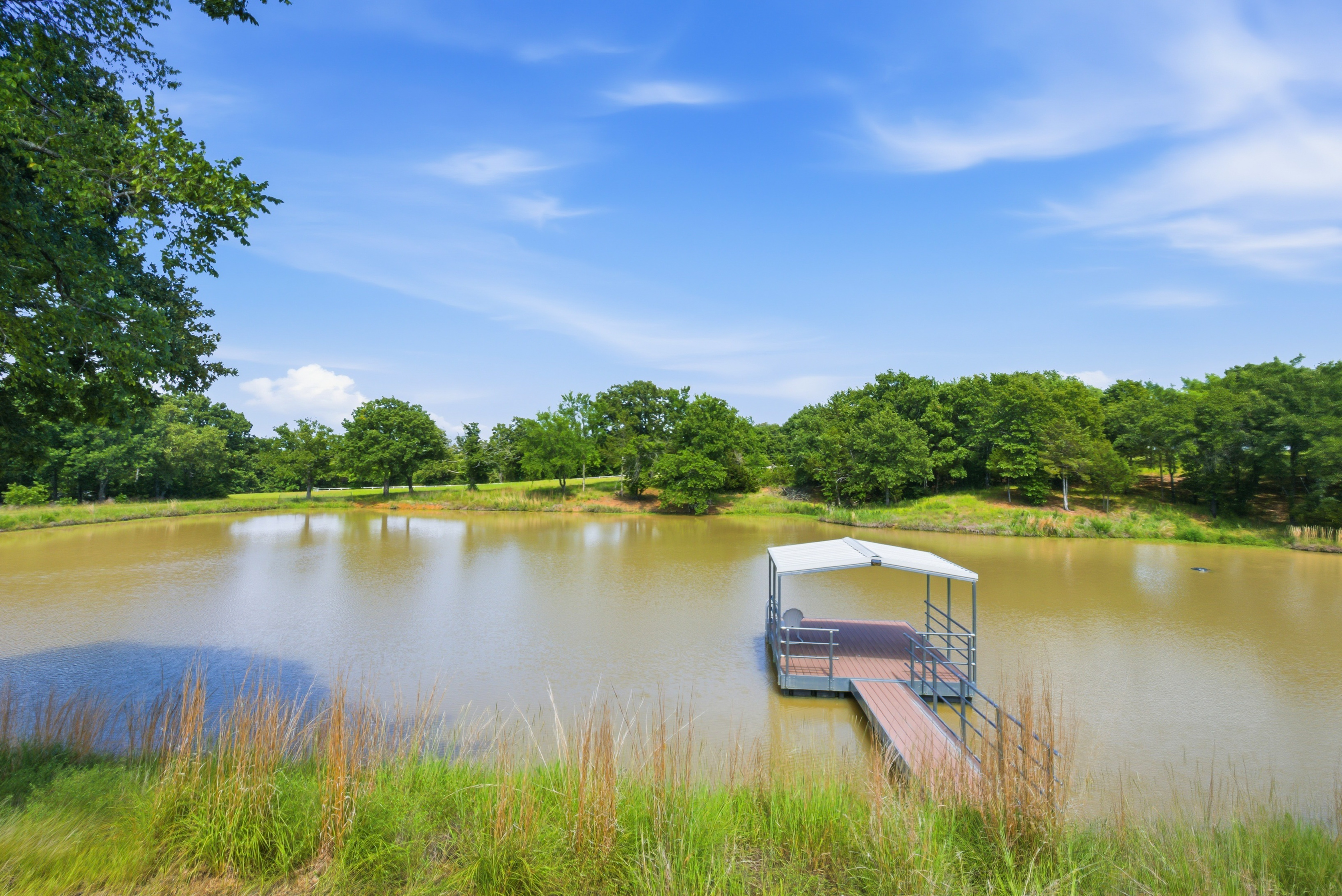 Metal-framed covered dock extending over the private pond, surrounded by lush green hills and mature oak trees