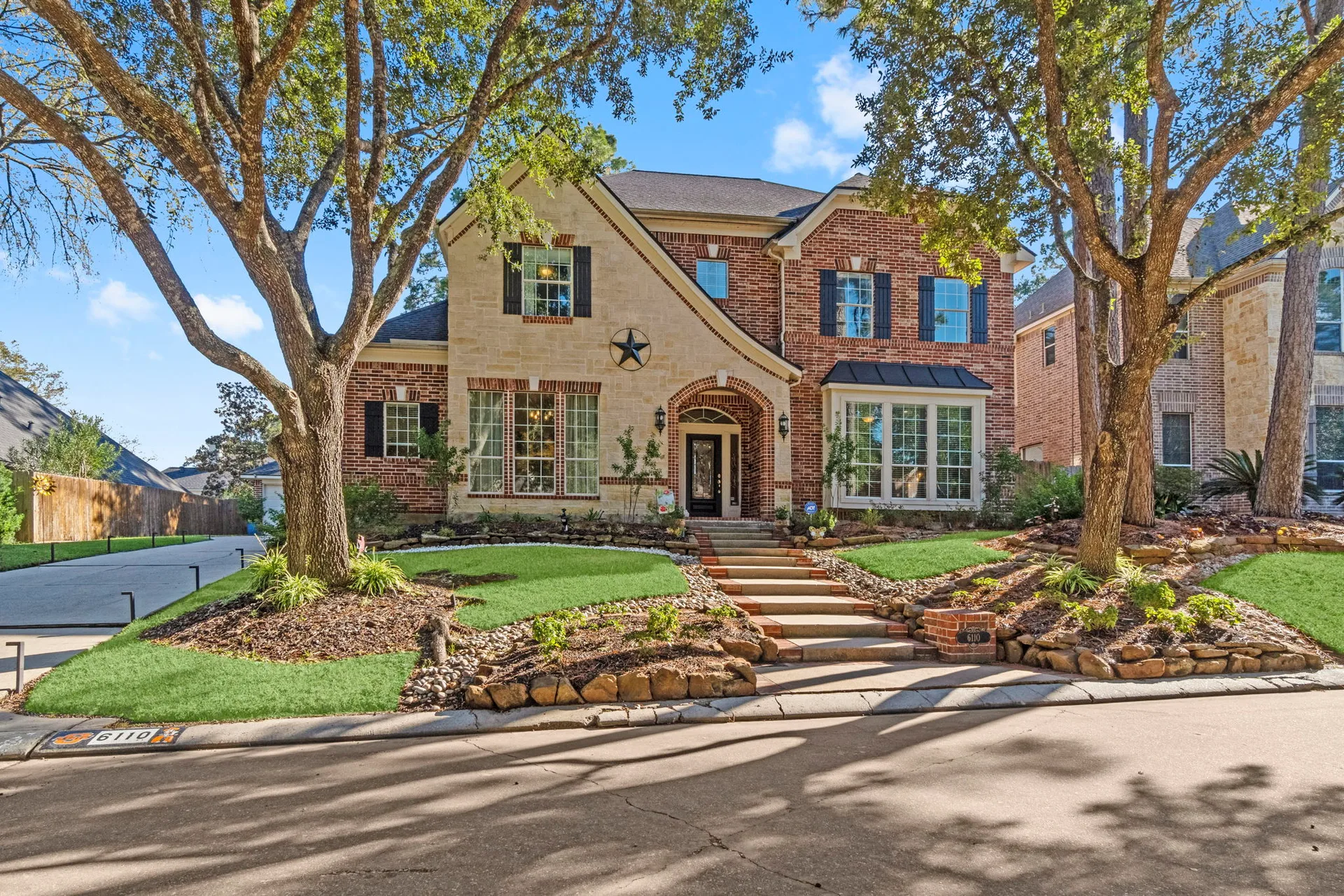 Front exterior of 6110 Peachtree Hill Court showing traditional brick and stone facade with mature oak trees under a clear blue sky