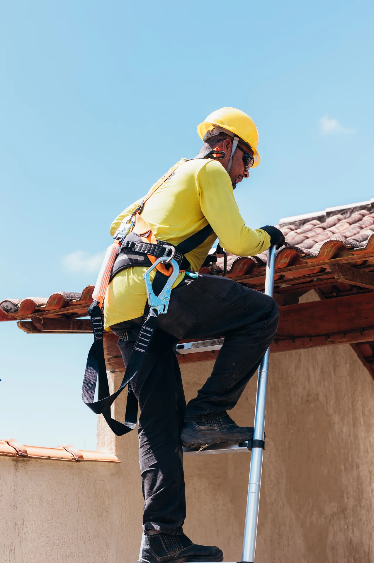 A contractor wearing a yellow hard hat and safety harness climbing a ladder to inspect a residential roof