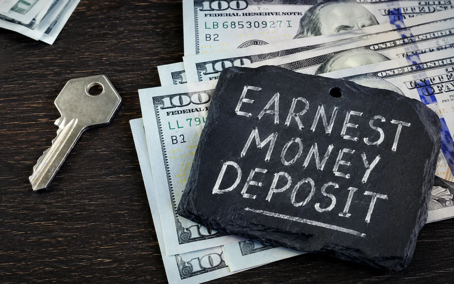 US hundred dollar bills and a house key next to an Earnest Money Deposit stone plaque