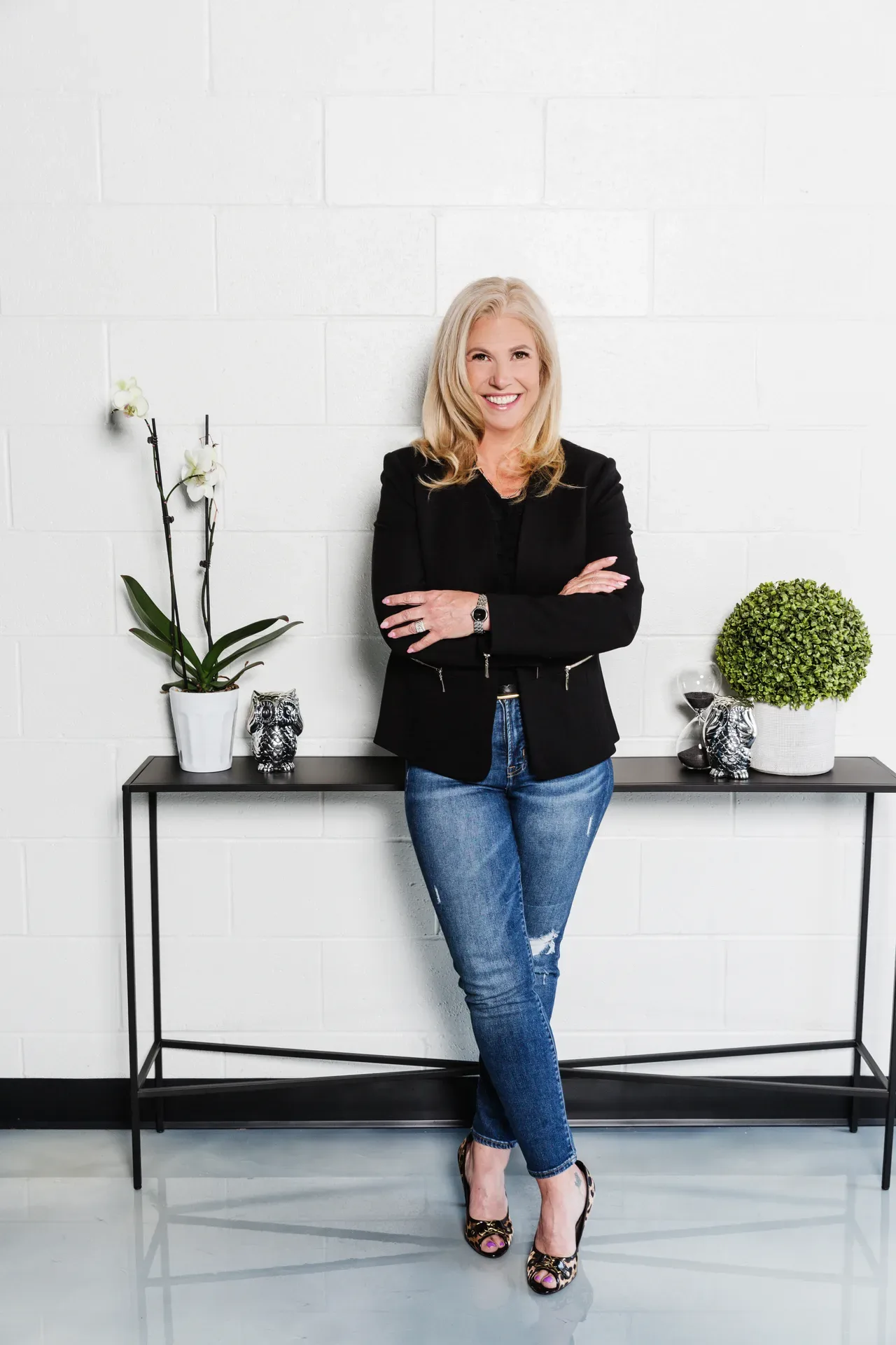Lee Lander in a black blazer and heels, arms crossed confidently in front of a white brick wall