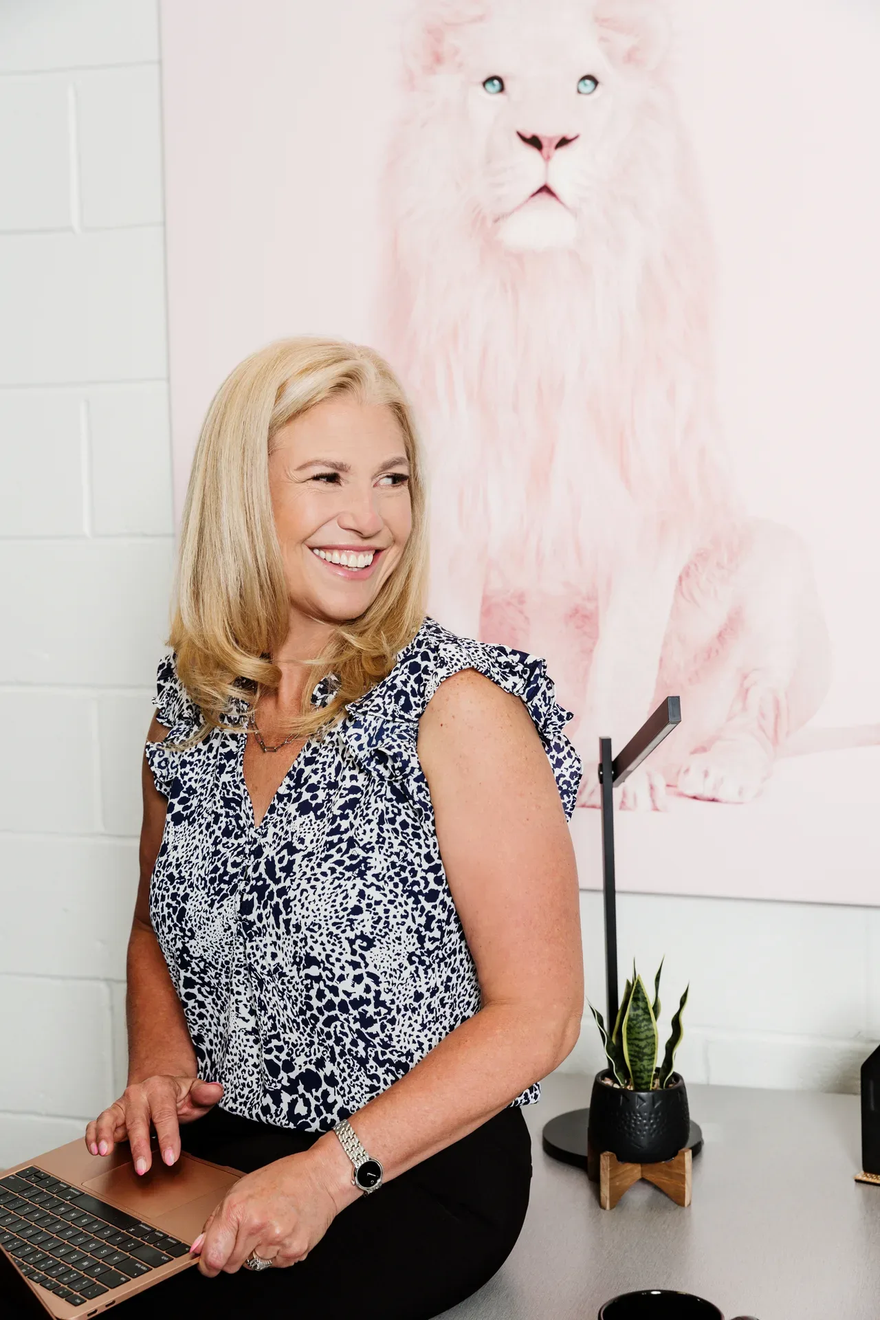 Lee Lander smiling at her desk with a laptop, with a pink lion artwork on the white brick wall behind her