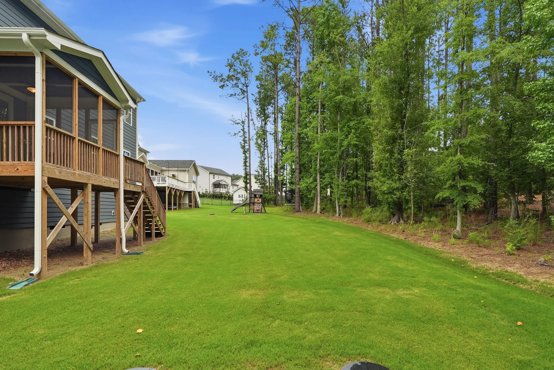 Rear elevation showing elevated screened-in deck with stairs, green lawn, and wooded privacy