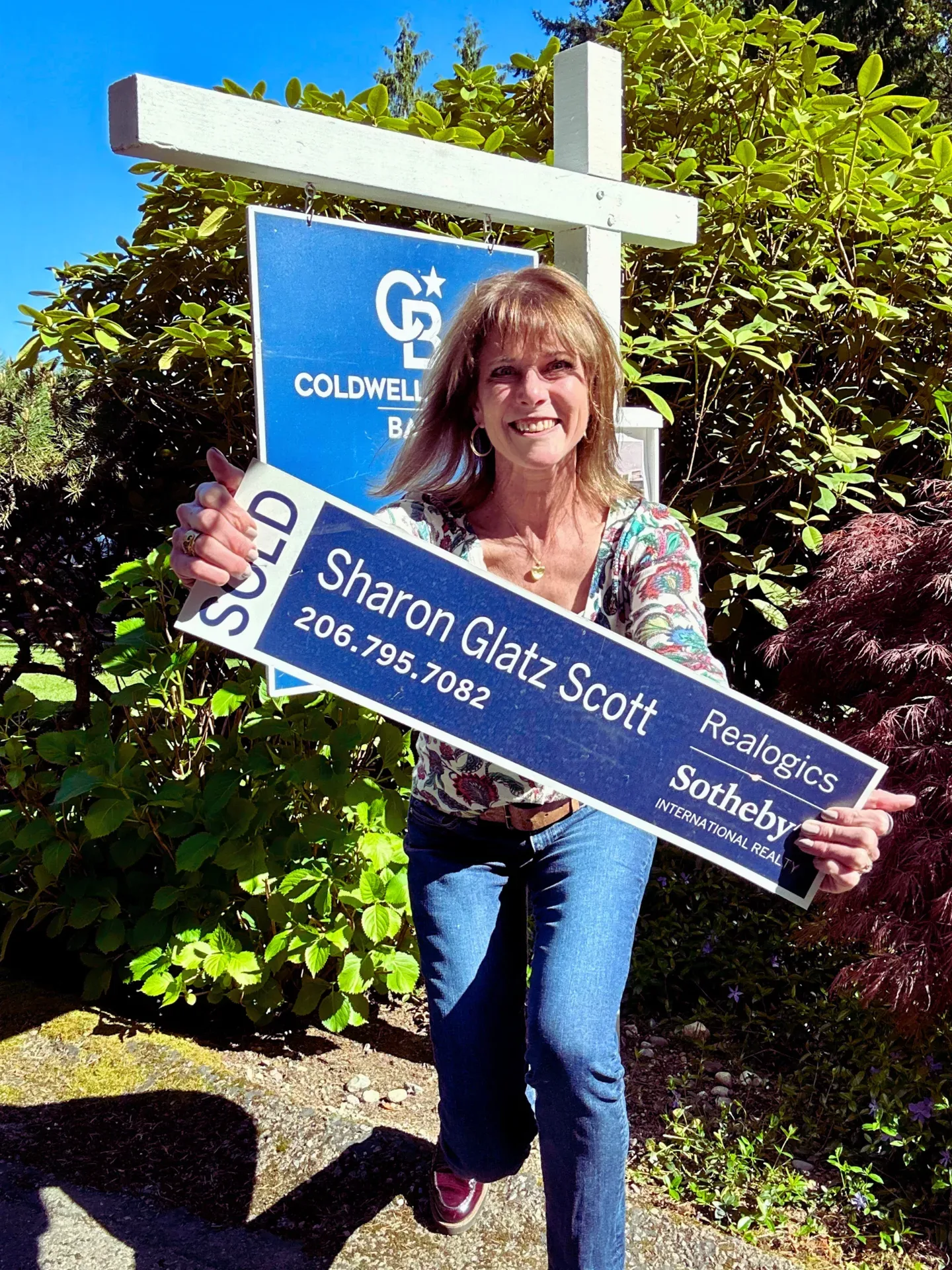 Sharon Glatz Scott holds a Sold sign in front of a real estate post outdoors