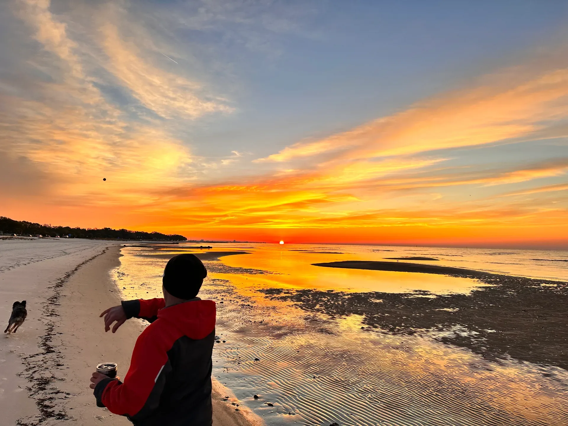 A person stands on a sandy beach throwing a ball for a dog during a vibrant orange sunset on the Mississippi Gulf Coast
