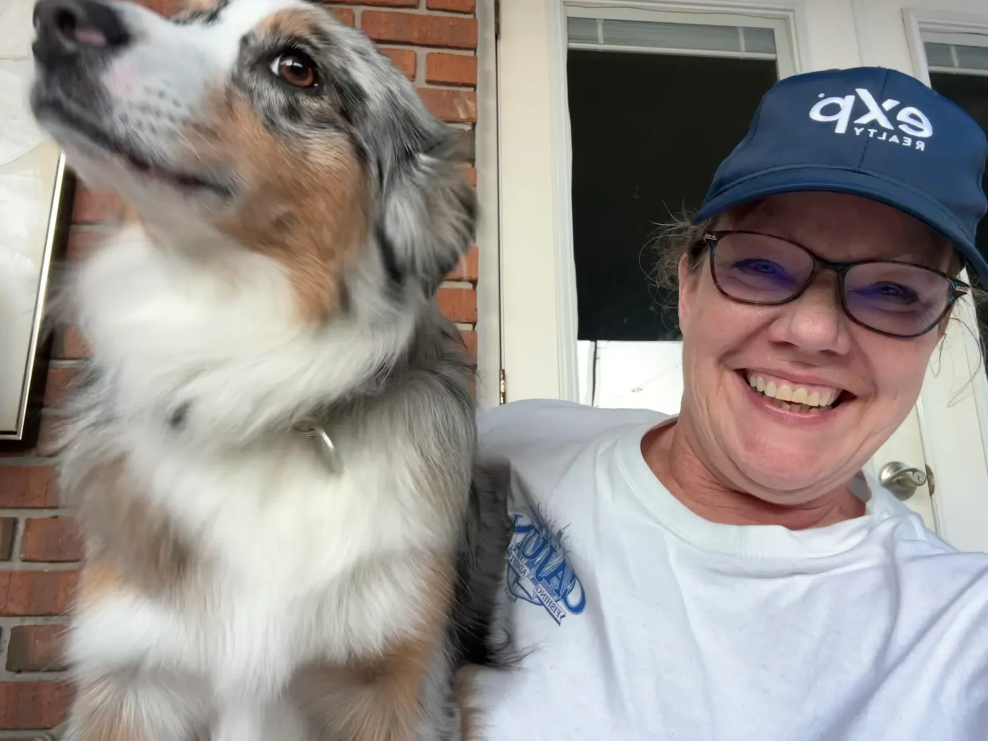 Judy Atherton wearing an eXp Realty cap while posing outdoors next to her tricolor Australian Shepherd