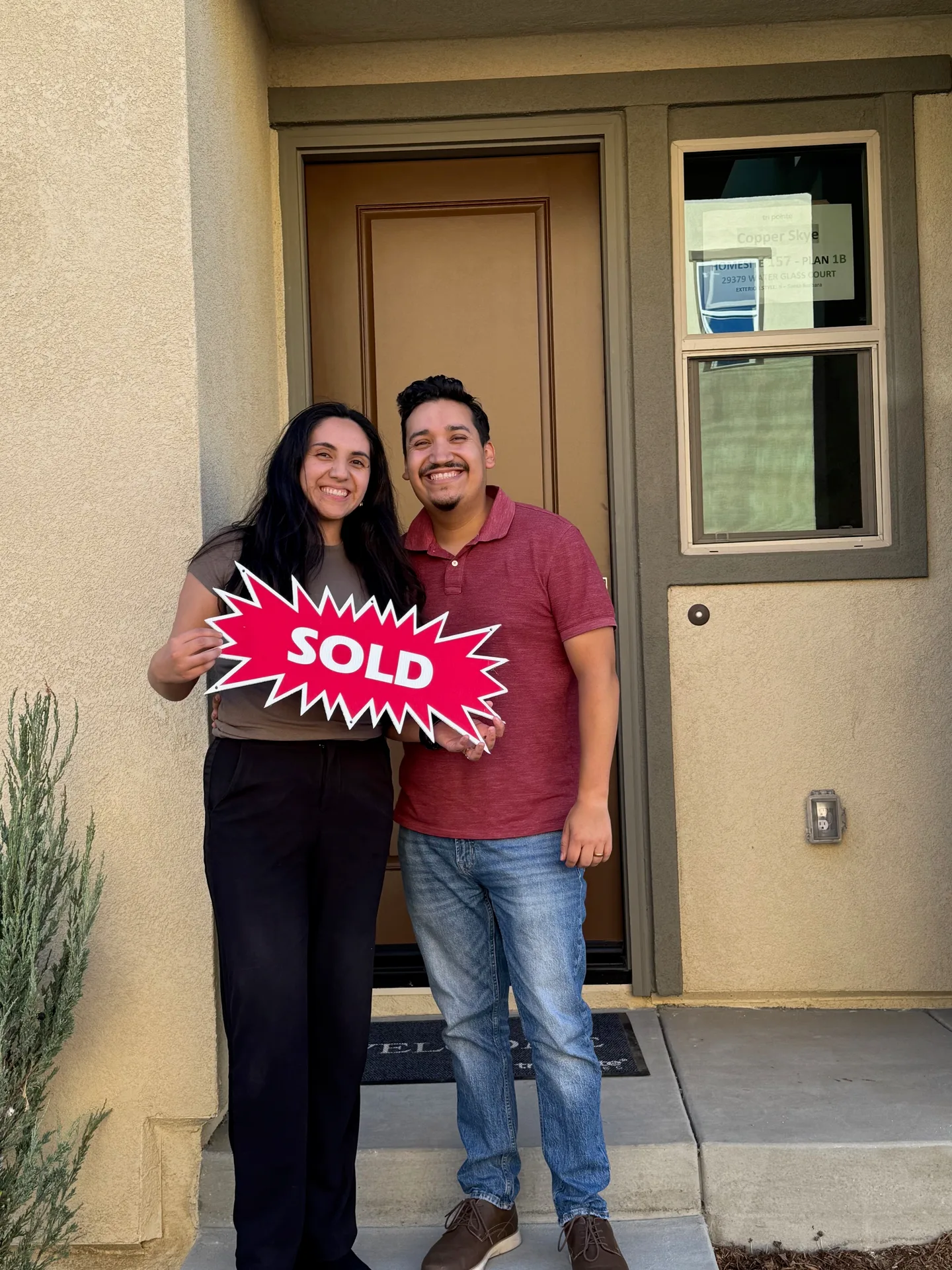 Isaac and Katie standing at the front entry of their new home with a sold sign, smiling on closing day