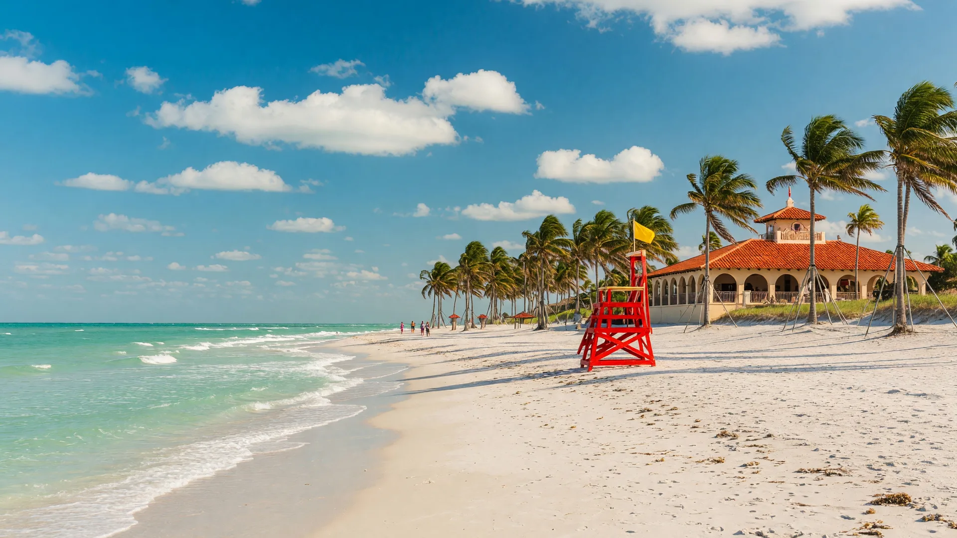 Boca Raton beach in Florida — wide expanse of white sand, turquoise Atlantic Ocean, blue sky with scattered clouds, palm trees, and the Spanish-style pavilion at Red Reef Park