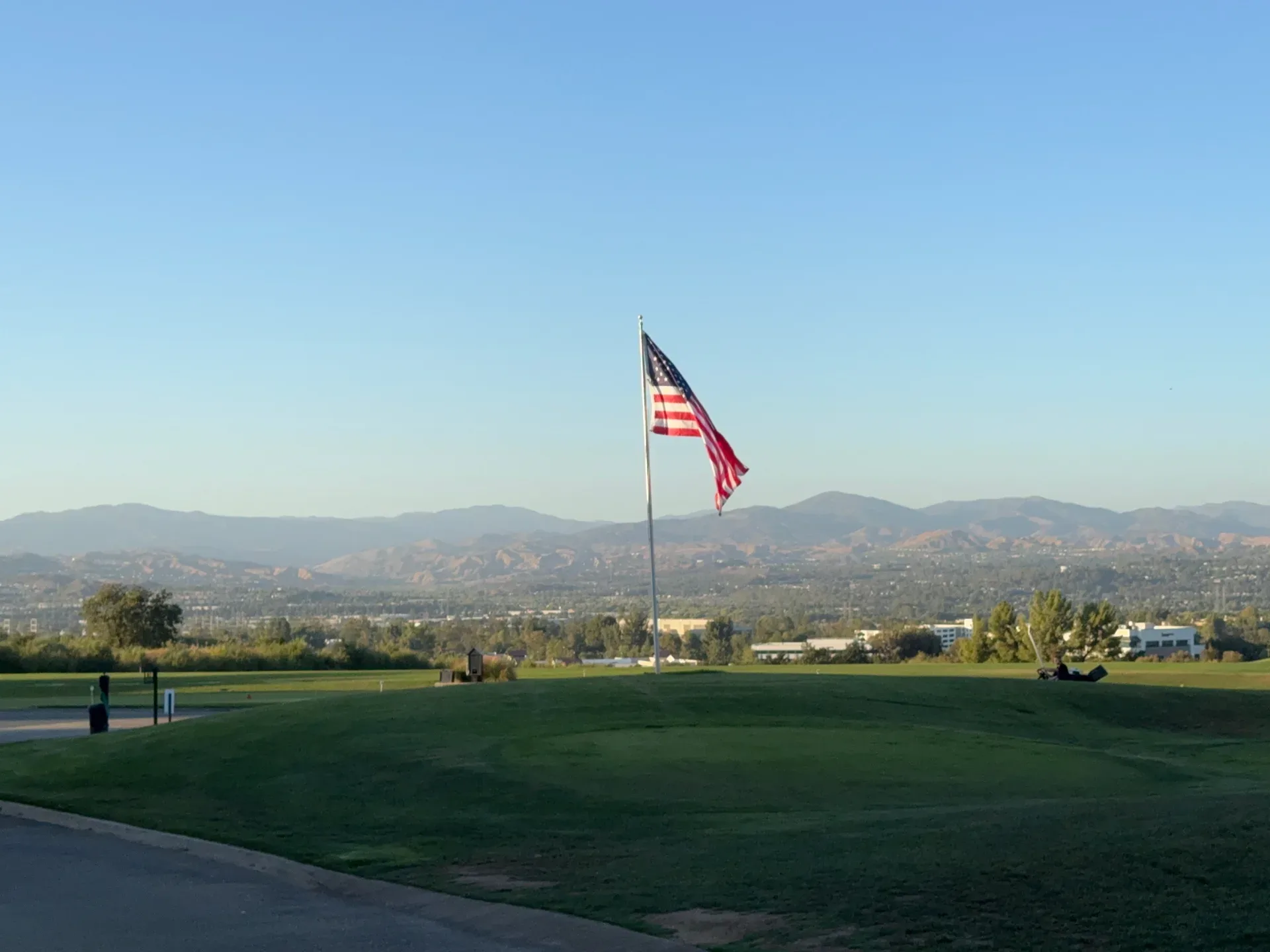An American flag flies above a green field with rolling mountains under a clear blue sky