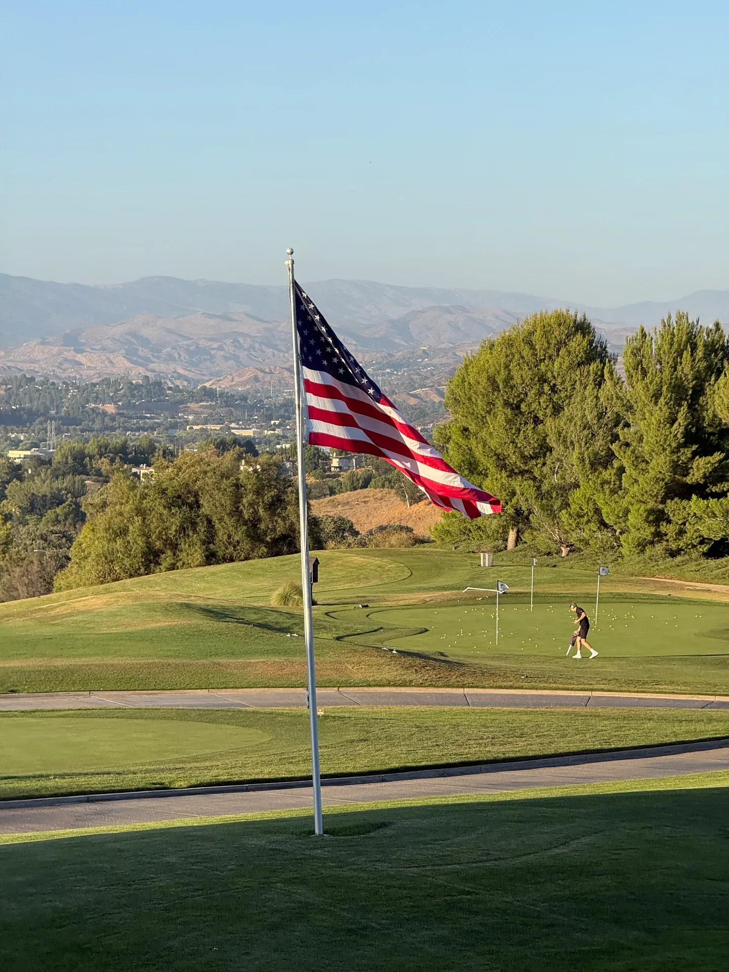 An American flag flies over a green golf course with mountains in the background