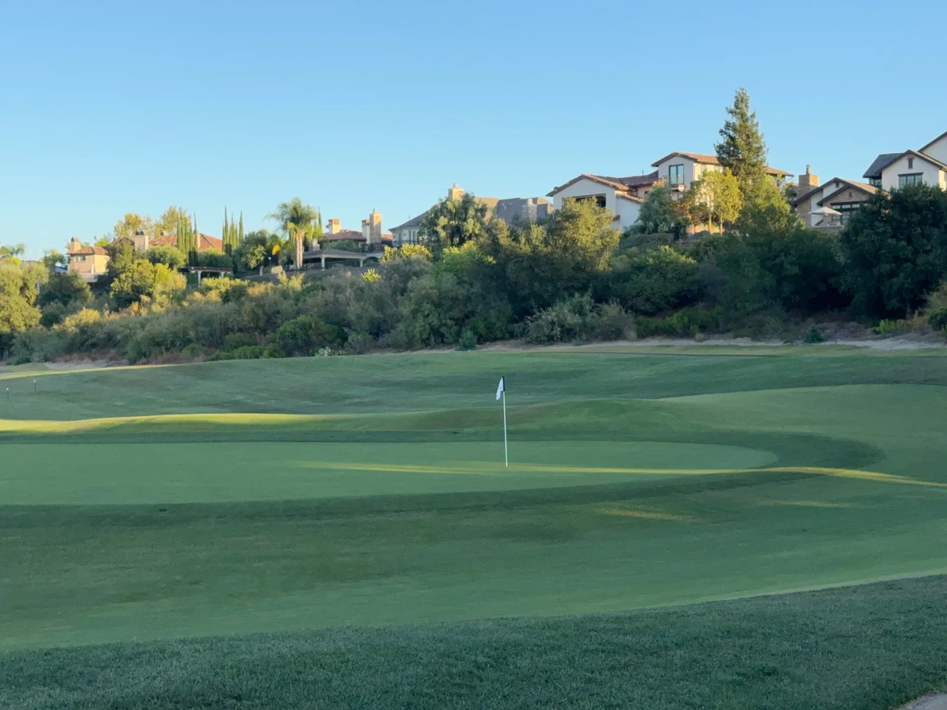 A well-maintained golf course green with houses visible in the background under a clear sky, showcasing Southern California living