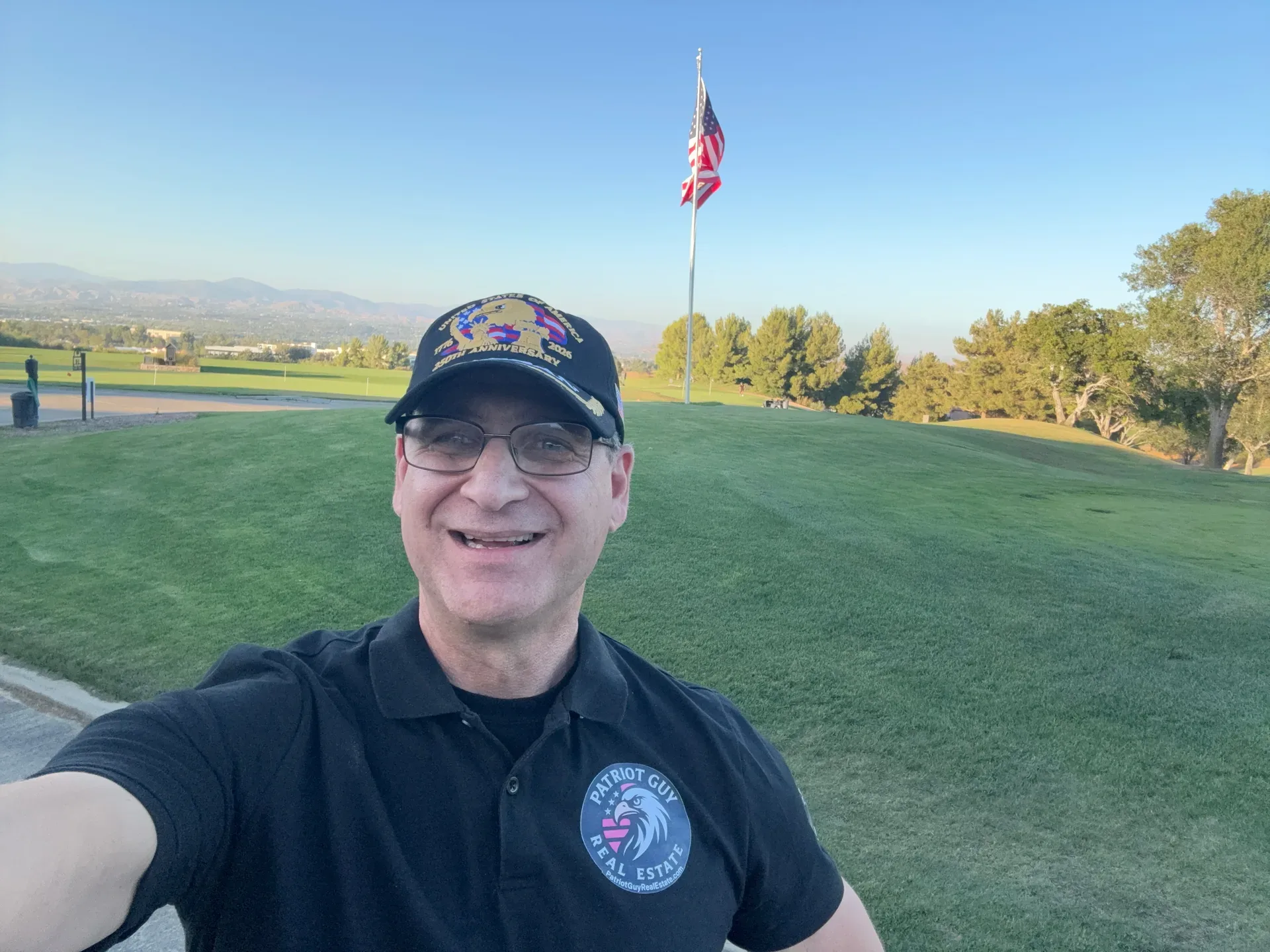 Sam Silver takes a selfie outdoors in a polo shirt and ball cap, standing in front of a green field with an American flag flying in the background