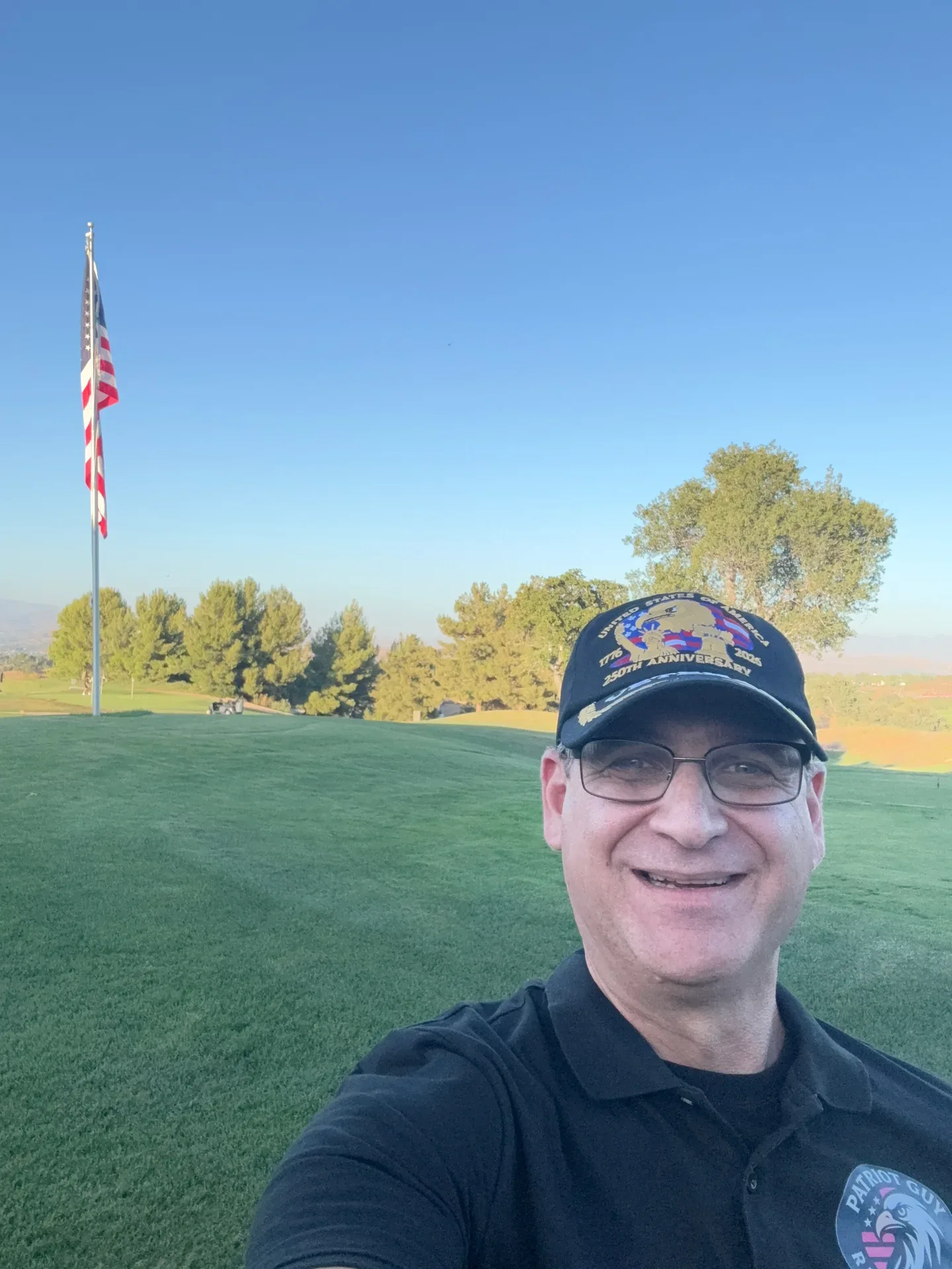 Sam Silver smiling outdoors on a sunny day at a golf course with an American flag flying in the background, enjoying time with the community